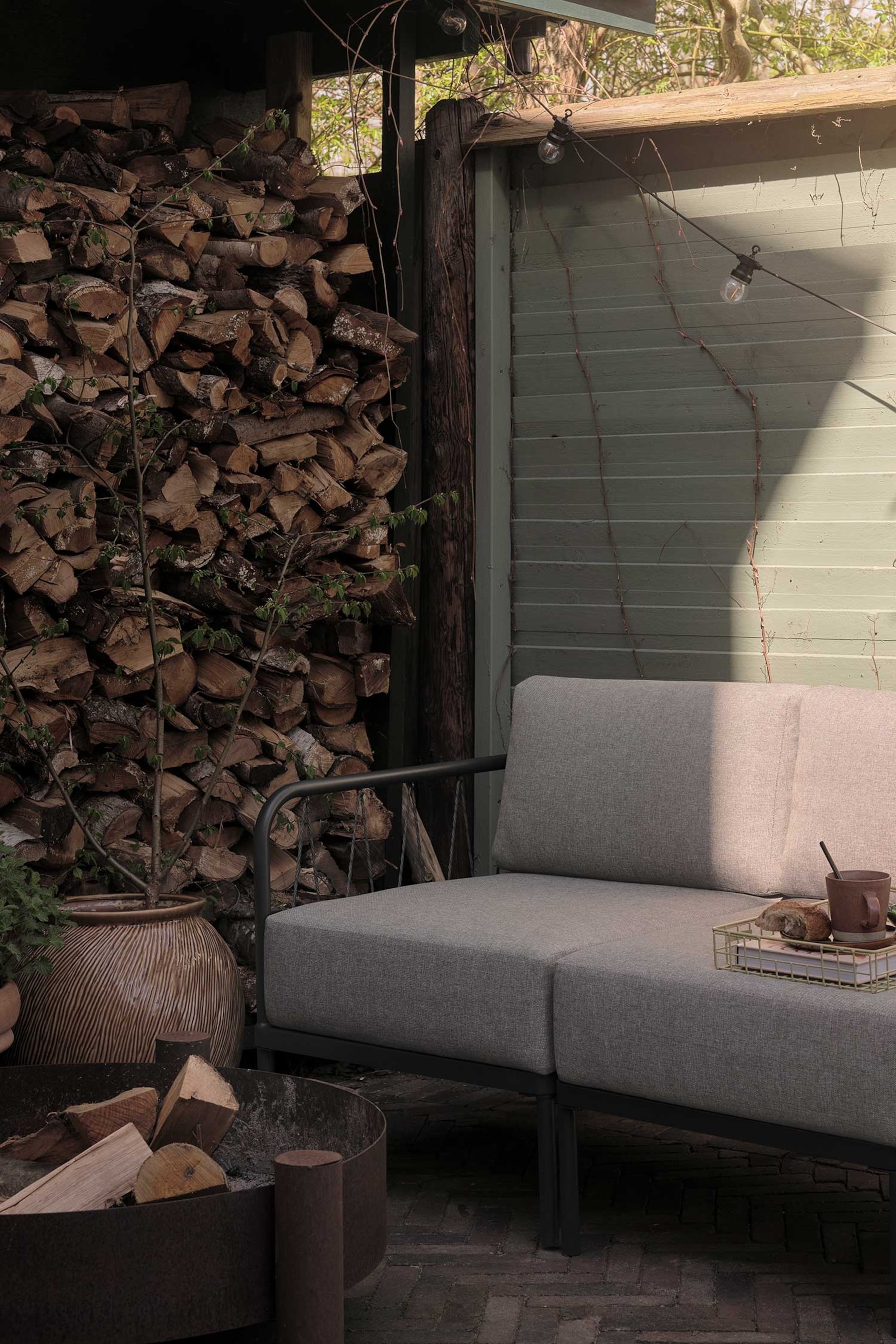 Größeres Bild anzeigen: Ein Outdoor-Sofa mit einem modularen Design, das mit hellgrauem Stoffbezug und einem dunklen Metallrahmen ausgestattet ist, der leicht zum Betrachter geneigt ist. Es besteht aus zwei Sitz- und zwei Rückenkissen, wobei ein hellgoldenes Drahttablett mit einer Tasse und einem Brot auf dem rechten Sitzkissen ruht.