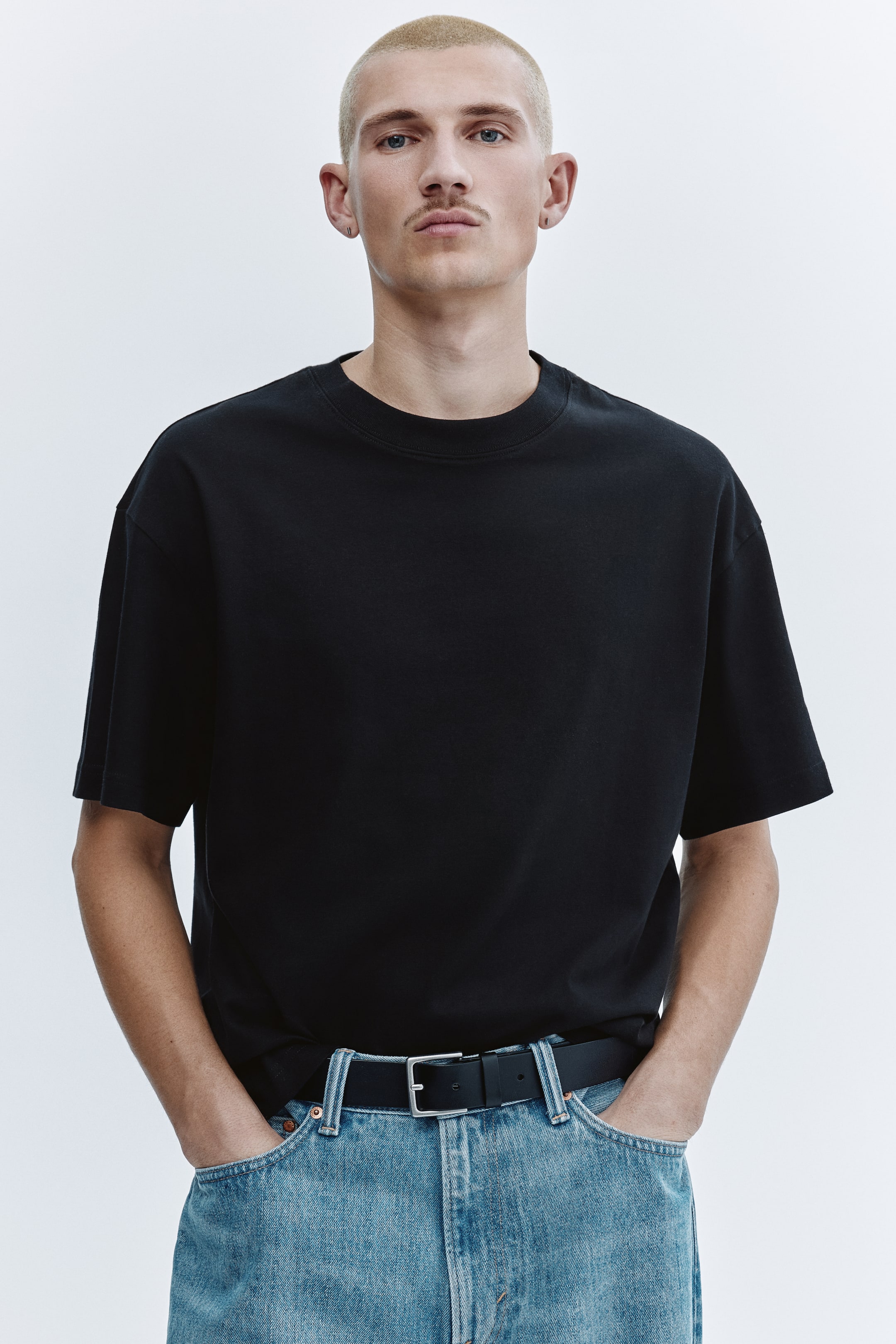 הצגת תמונה גדולה יותר: חולצת טי שחורה בצווארון עגול במידת אוברסייז, עם כתפיים יורדות ושרוולים קצרים ורחבים. החולצה מוצגת מכנוסה לתוך ג'ינס תכלת בהיר, והבד חלק במראה מאט.