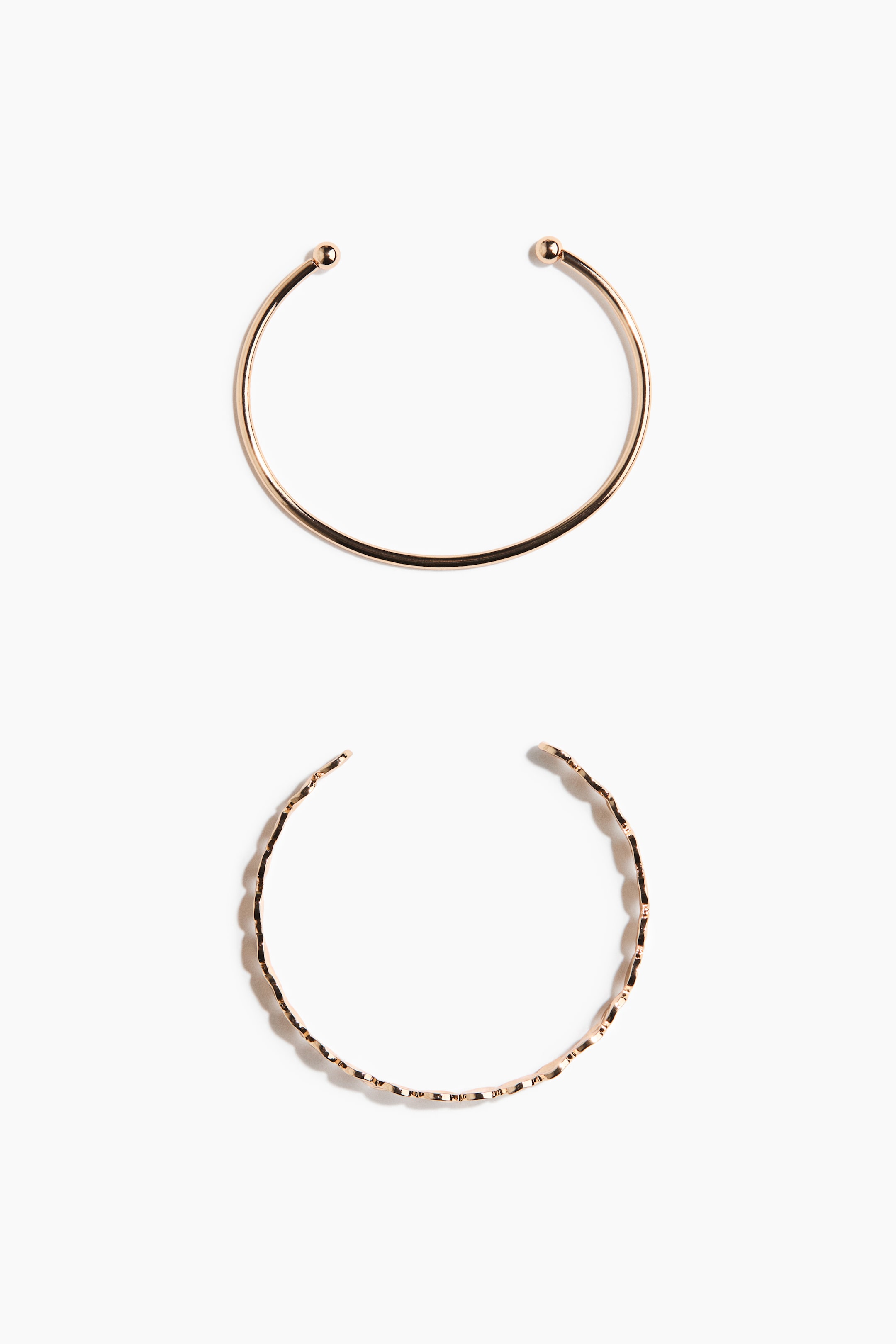 View larger image: Two gold-toned open-ended bangles are lying flat, showcasing distinct designs. The top bangle is featuring a smooth, circular band with spherical beads capping each end, while the bottom bangle is displaying a hammered, wavy band with plain ends.