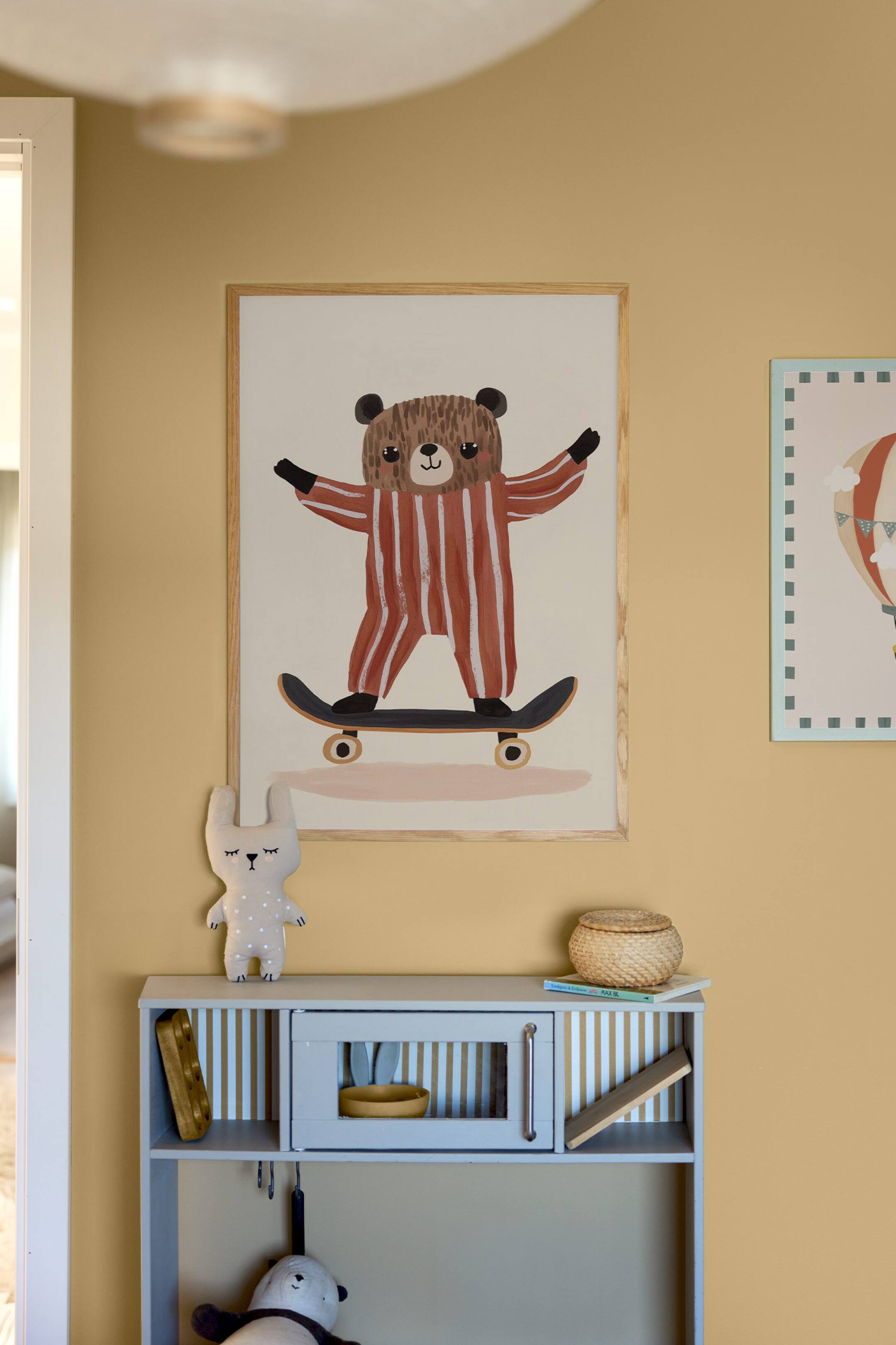 Grotere afbeelding bekijken: Een rechthoekige ingelijste kunstprint toont een illustratie van een bruine beer die rood-wit gestreepte pyjama draagt terwijl hij op een skateboard rijdt, tegen een gebroken witte achtergrond. De print is omlijst met een licht naturel houten frame en hangt plat aan een muur.