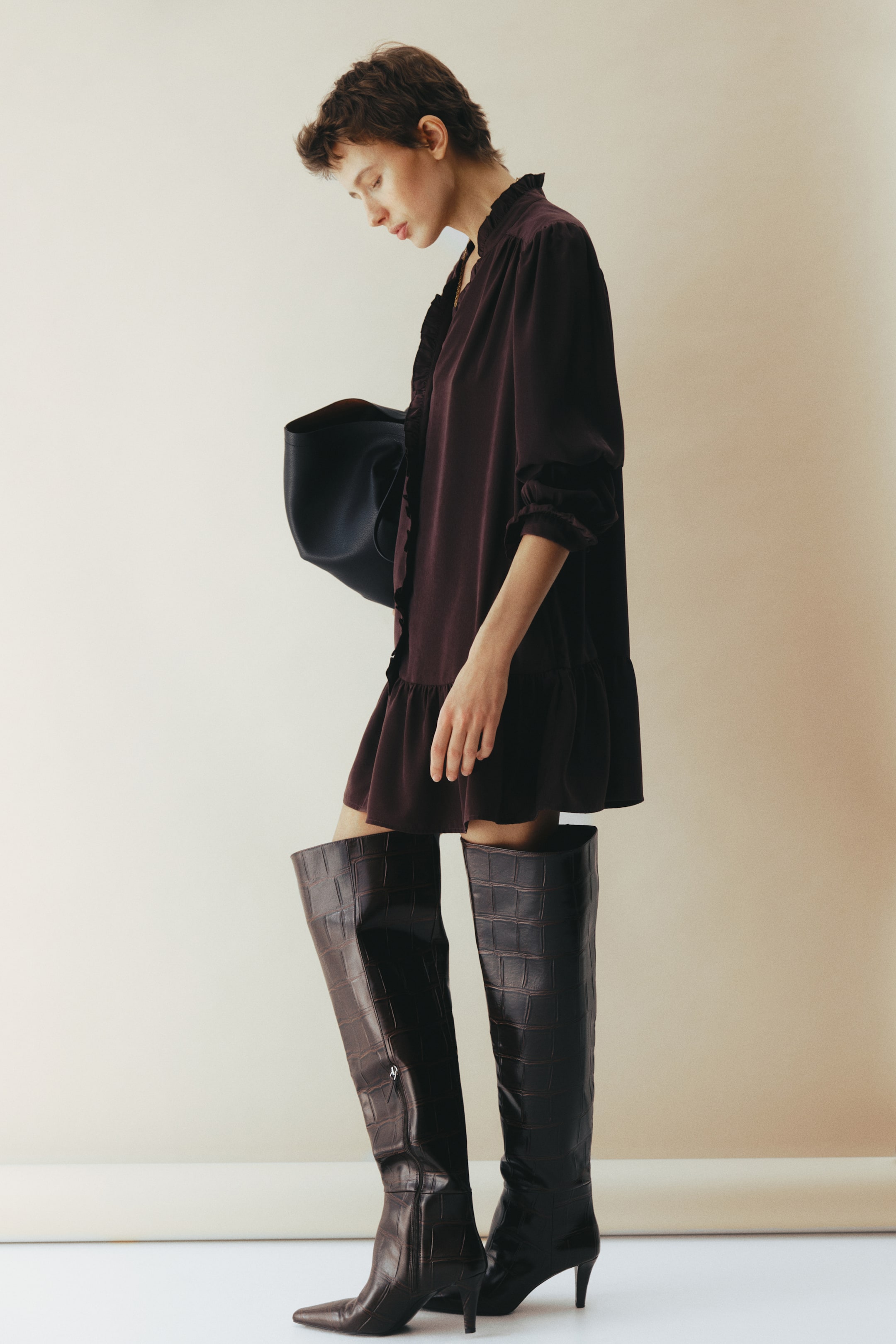 View larger image: A short, oversized dark reddish-brown dress, featuring a V-neckline with frill trim, buttons down the front, forward-facing shoulder seams with gathers, long balloon sleeves with frilled cuffs, and a tiered hem, is worn by the model. The model is also wearing dark knee-high embossed boots and carrying a dark bag.