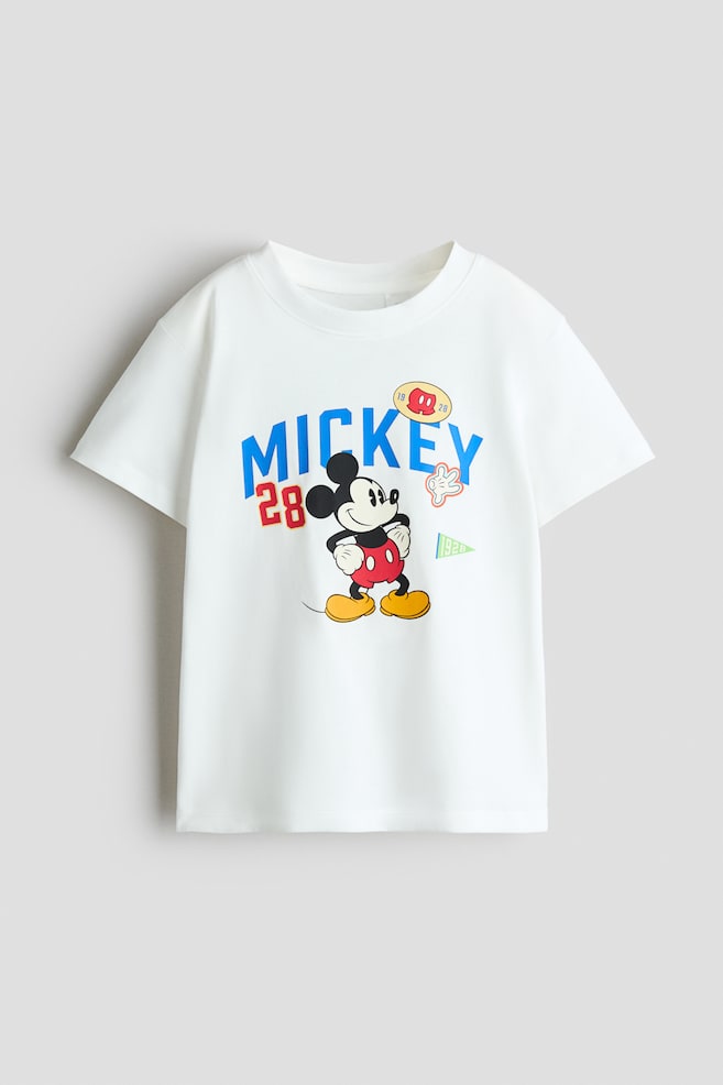 A white cotton jersey t-shirt is featuring a printed Mickey Mouse graphic on the front, accented by the word "MICKEY" in blue arched lettering and the number "28" in red. The t-shirt is showcasing a round, rib-trimmed neckline and short sleeves with gently dropped shoulders. - 1