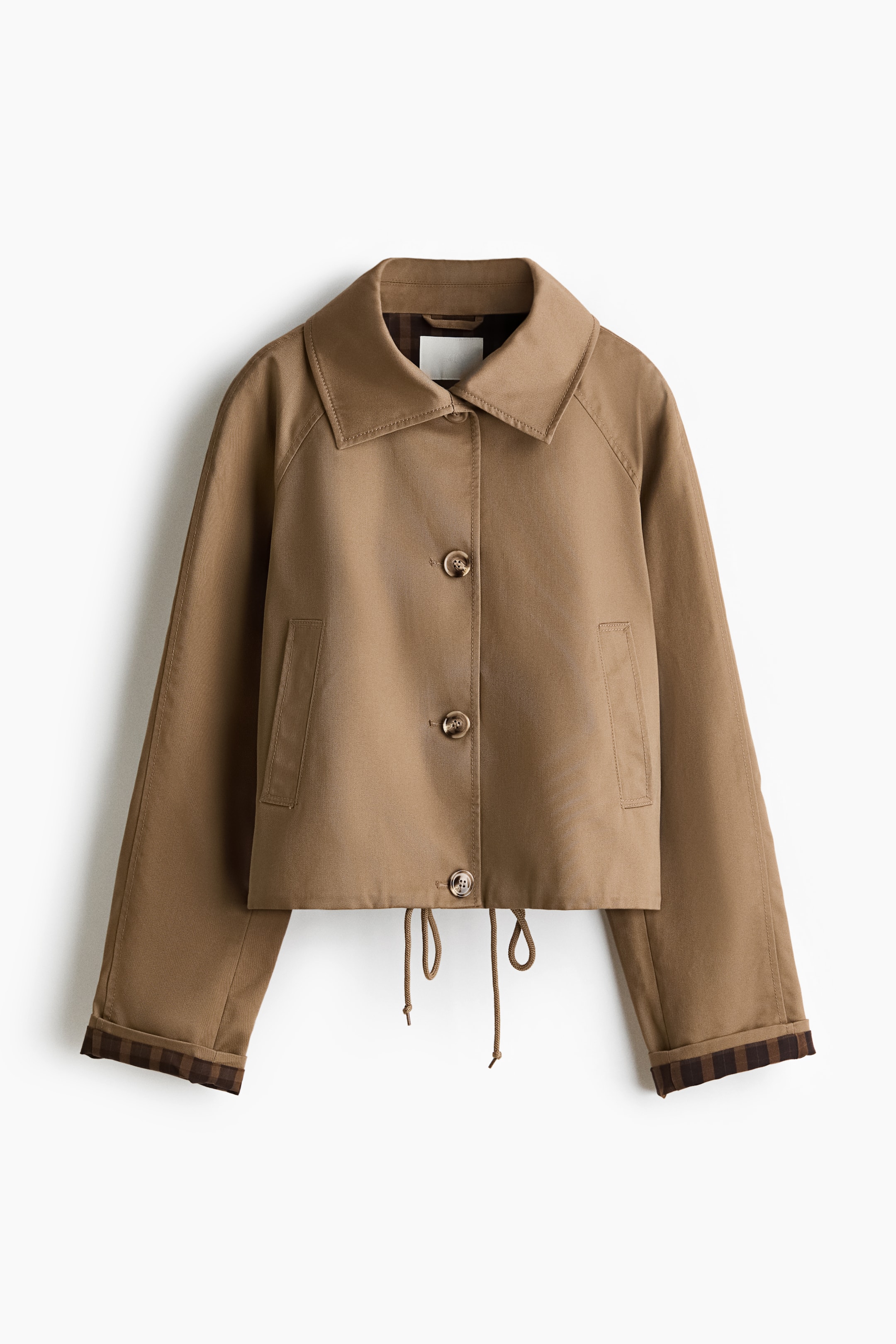 Visualizza immagine più grande: Una giacca marrone è mostrata frontalmente, con colletto ampio, chiusura frontale con quattro bottoni marroni e maniche raglan. Presenta polsini risvoltati che rivelano una fodera a quadri, due tasche a filetto orizzontali e un cordoncino sull'orlo corto.