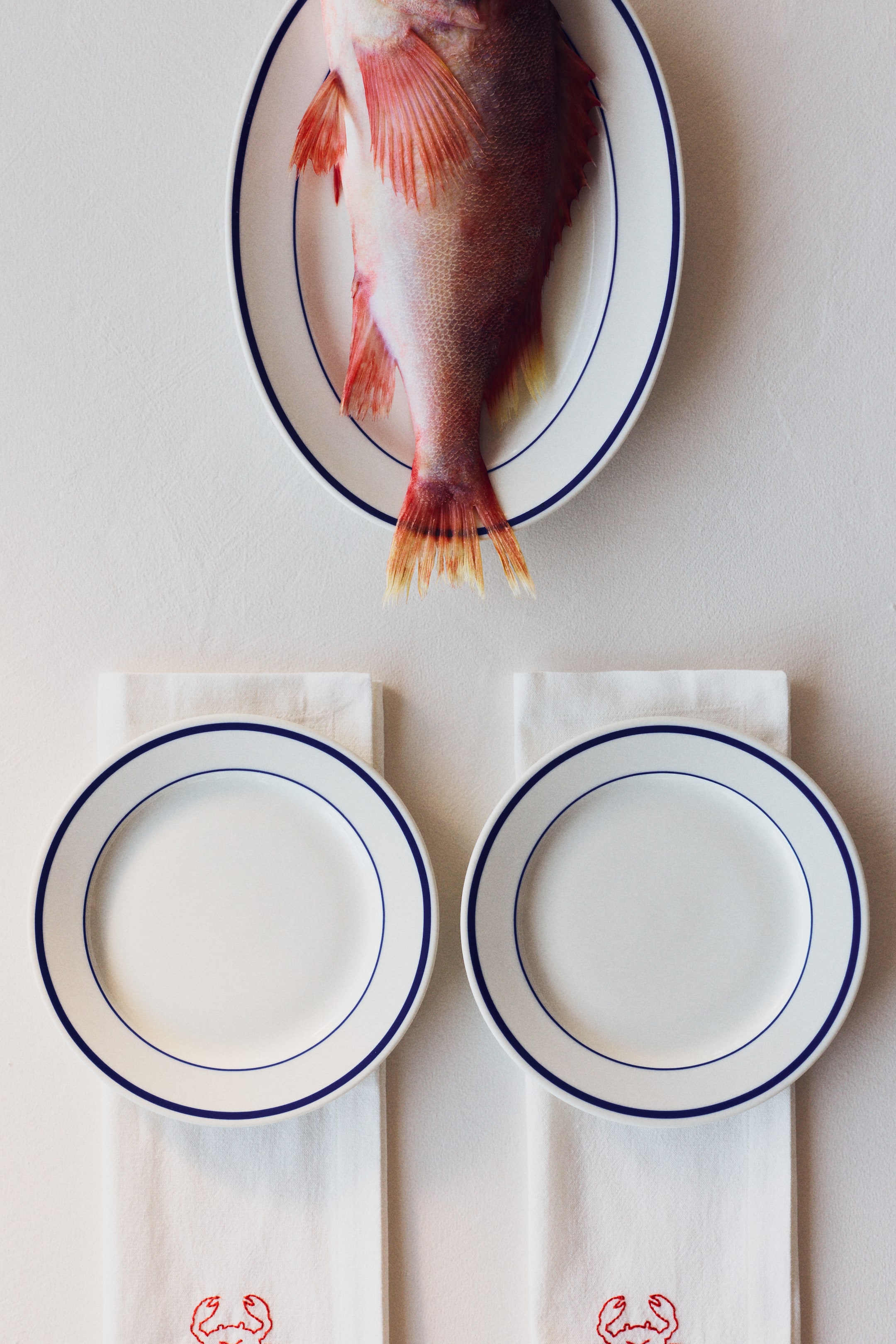 View larger image: A whole red fish is laid flat on an oval white plate with a blue rim, its tail pointing downwards. Below, two round white plates with blue rims are placed on folded white napkins, each adorned with a red crab embroidery.