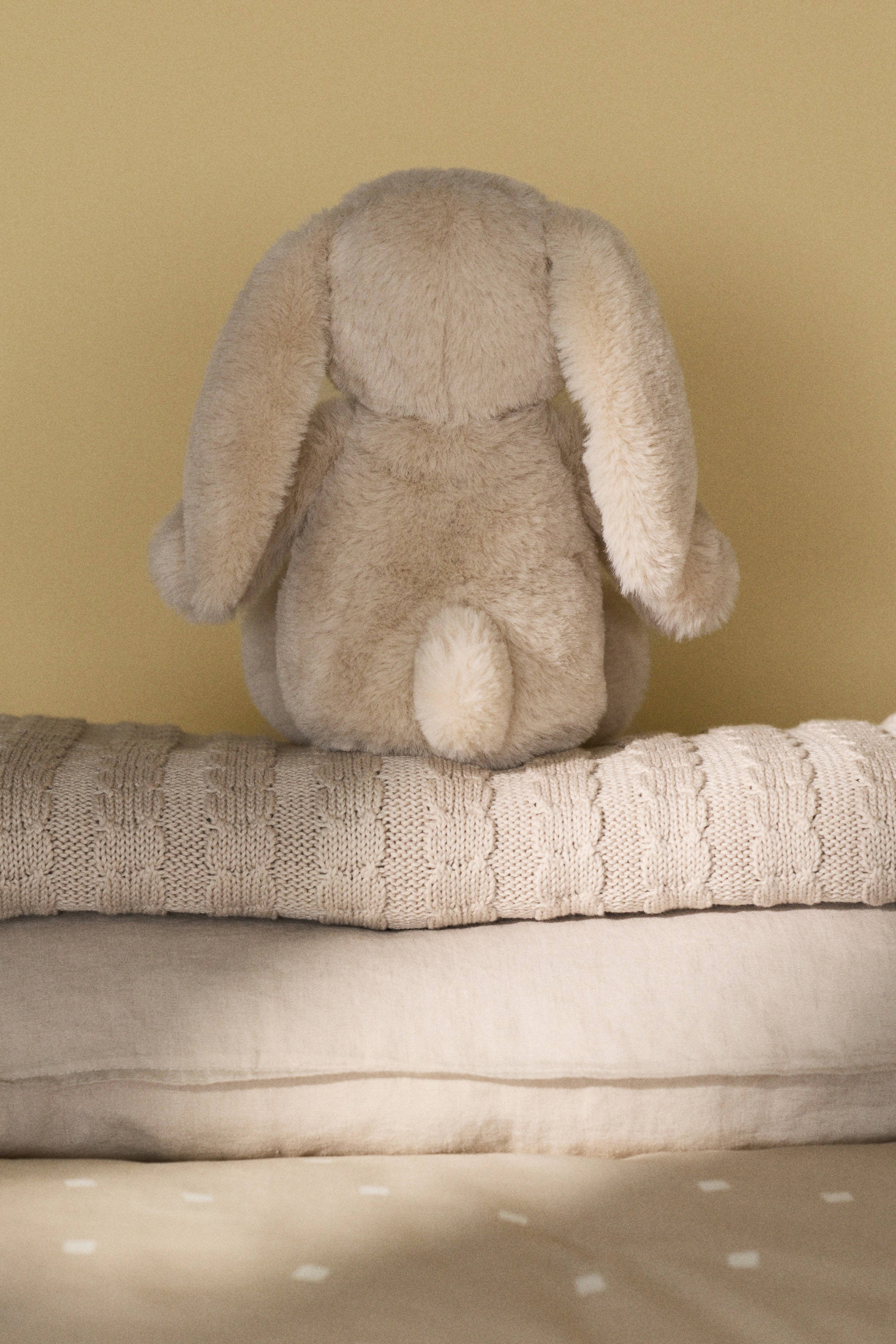 View larger image: Plush toy crafted from soft pile in the form of a bunny, showcasing adorable, embroidered facial features. Currently positioned facing away, it highlights long, floppy ears and a small, round, fluffy tail.