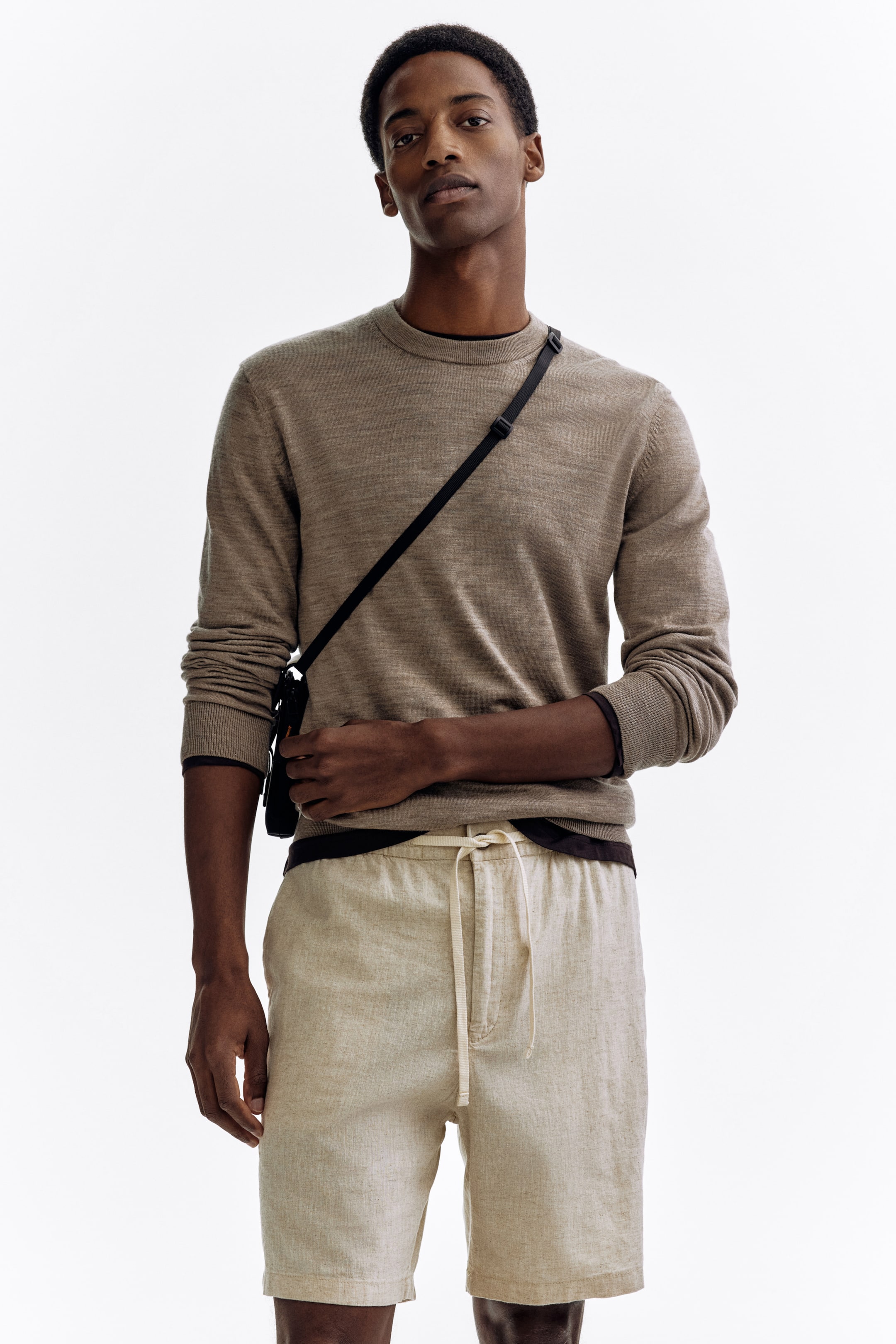 Agrandir l'image: Un pull à col rond marron clair présente des poignets côtelés et une couche sombre sous son ourlet côtelé, présenté de face. Il est associé à un short texturé en lin beige clair avec une taille à cordon de serrage.