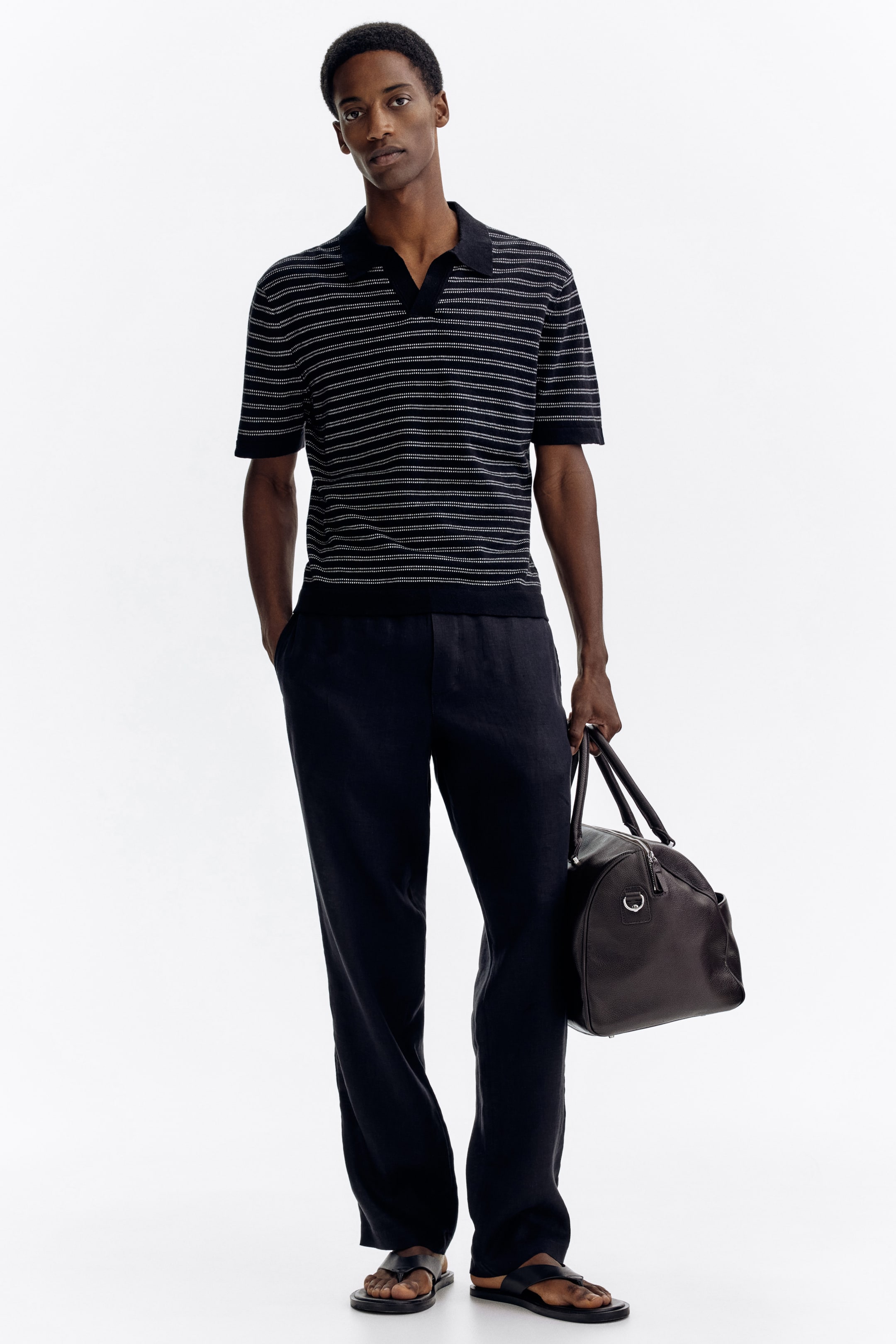 Grotere afbeelding bekijken: Een zwart gebreid poloshirt met korte mouwen, voorzien van dunne witte horizontale strepen en een open V-hals kraag, wordt gepresenteerd. Het is gestyled met donkerblauwe broek met een ontspannen pasvorm, zwarte leren sandalen met open teen, en een donkerbruine leren weekendtas met zilverkleurige details die gedragen wordt.