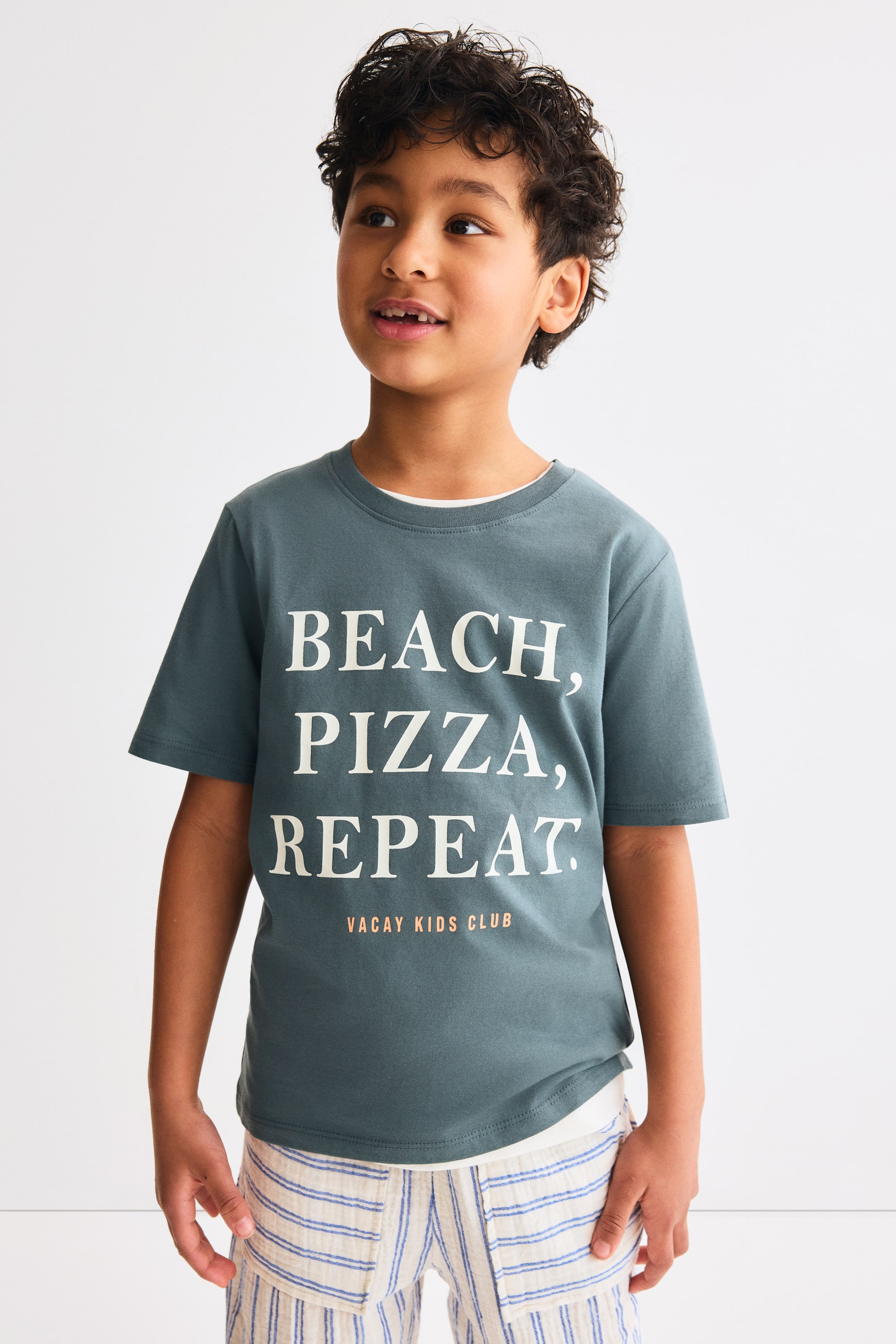 View larger image: A dark teal cotton jersey T-shirt features a white graphic print on the front that reads 'BEACH, PIZZA, REPEAT.' with 'VACAY KIDS CLUB' in orange text below. It is crafted with a ribbed crew neckline, dropped shoulders, and a straight-cut hem, with a white layer visible underneath.