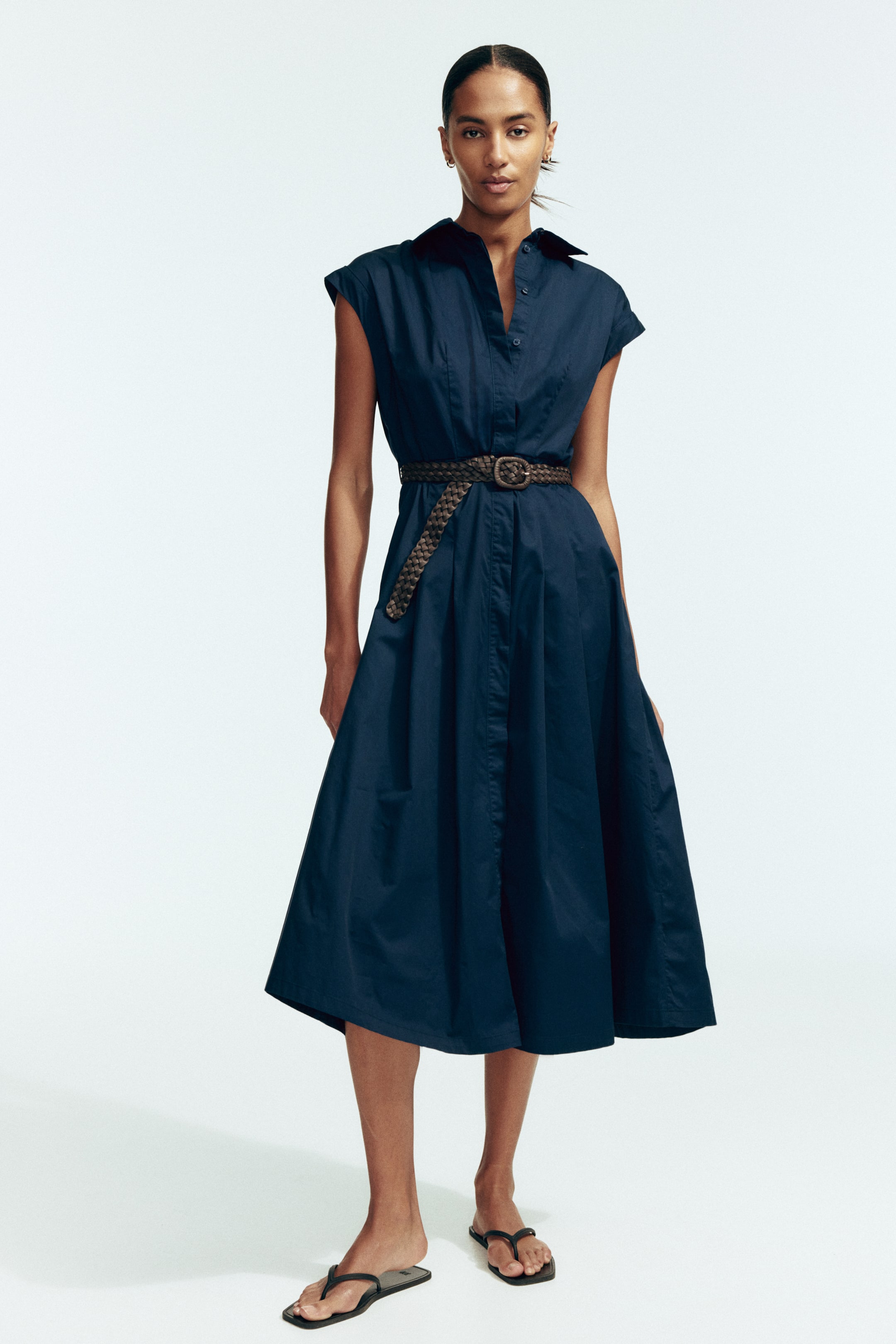 View larger image: A navy blue shirt dress is featuring a pointed collar, a button-down placket, and cap sleeves. The dress is cinched at the waist by a braided brown belt and is flowing into a pleated A-line midi skirt.
