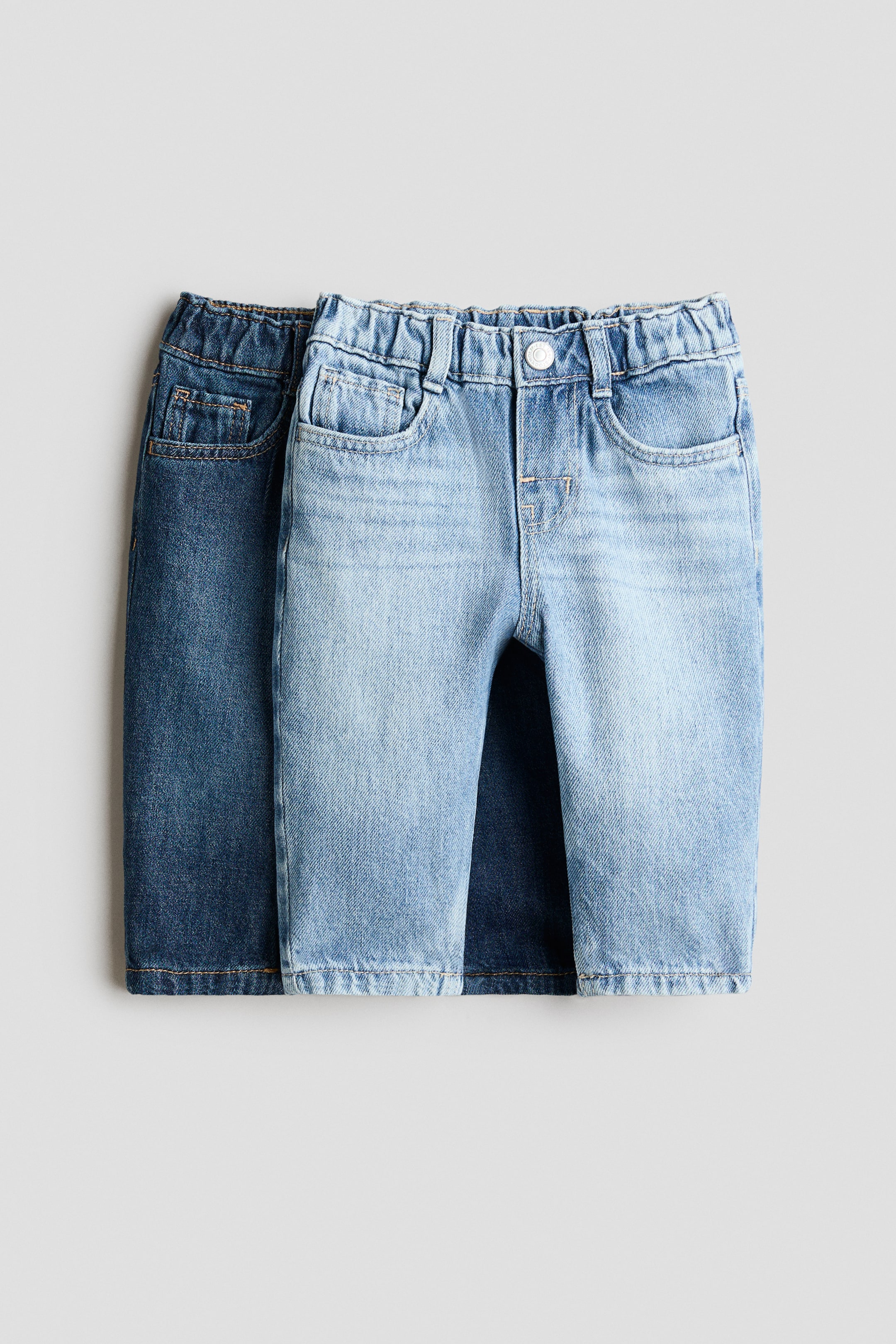 View larger image: Two pairs of jeans are displayed, with a light blue pair partially overlapping a darker blue pair. The light blue jeans are shown from the front, featuring an elastic waistband, belt loops, two front scoop pockets, and a white button closure.