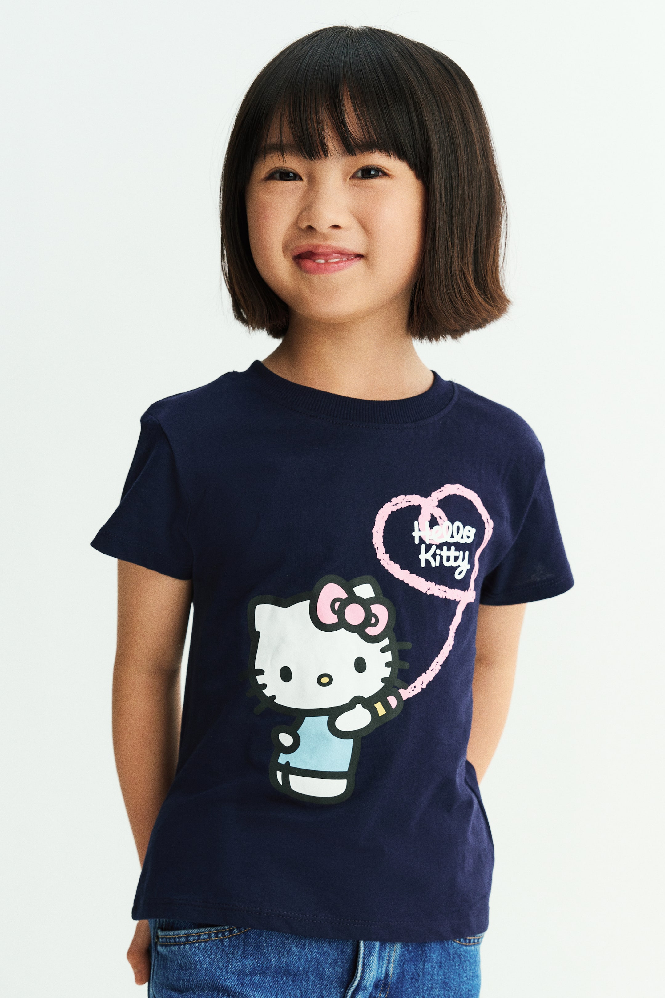 View larger image: A dark blue cotton jersey T-shirt featuring a Hello Kitty graphic print on the front is worn by the model. The T-shirt has a round, rib-trimmed neckline and short sleeves, and is styled with blue denim jeans.