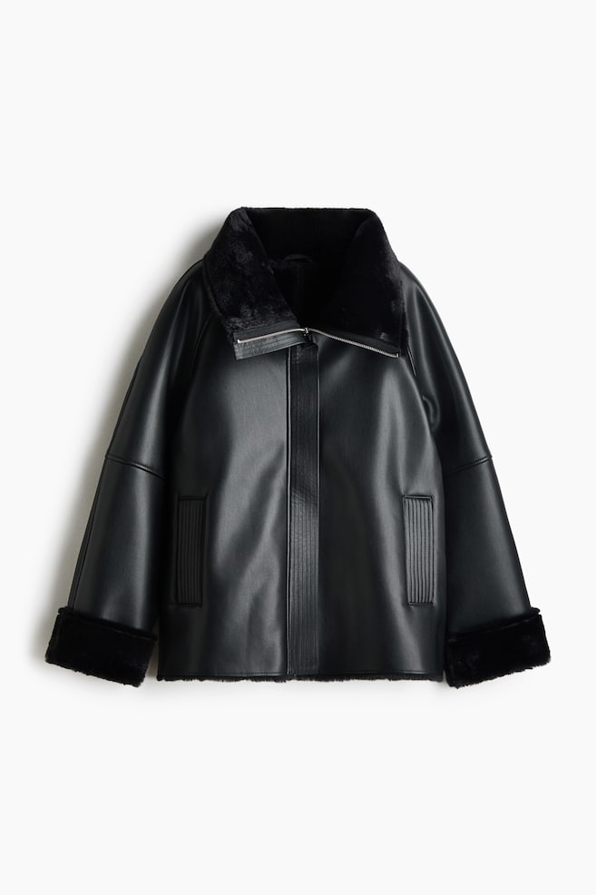 Se muestra una chaqueta de polipiel negra vista de frente, con un amplio cuello de pelo sintético negro y puños rematados a juego con pelo sintético. Incorpora un cierre asimétrico con cremallera cerca del cuello, una tapeta central cubierta y bolsillos de vivo verticales con detalle acanalado en cada lado. - 1