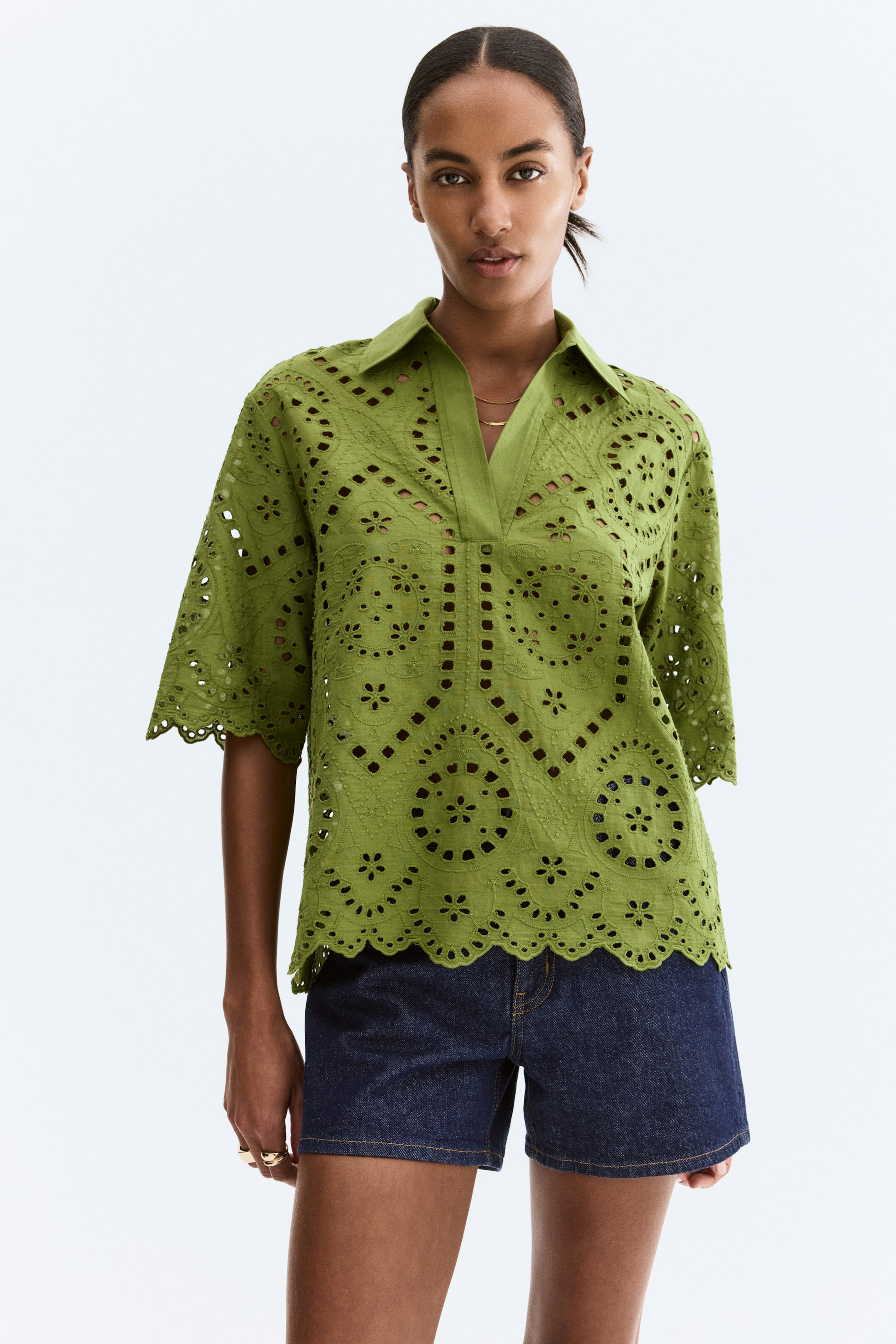Xem ảnh lớn hơn: Một chiếc áo màu xanh ô liu broderie anglaise đang hướng mặt trước, có đường viền cổ chữ V và cổ áo trải rộng. Nó nổi bật với các họa tiết thêu cắt tinh xảo trên toàn thân và tay áo dài đến khuỷu, với các cạnh được viền hình vỏ sò. Đường viền dưới của áo cũng được viền hình vỏ sò, và một chiếc vòng cổ xích vàng có thể thấy qua cổ chữ V.