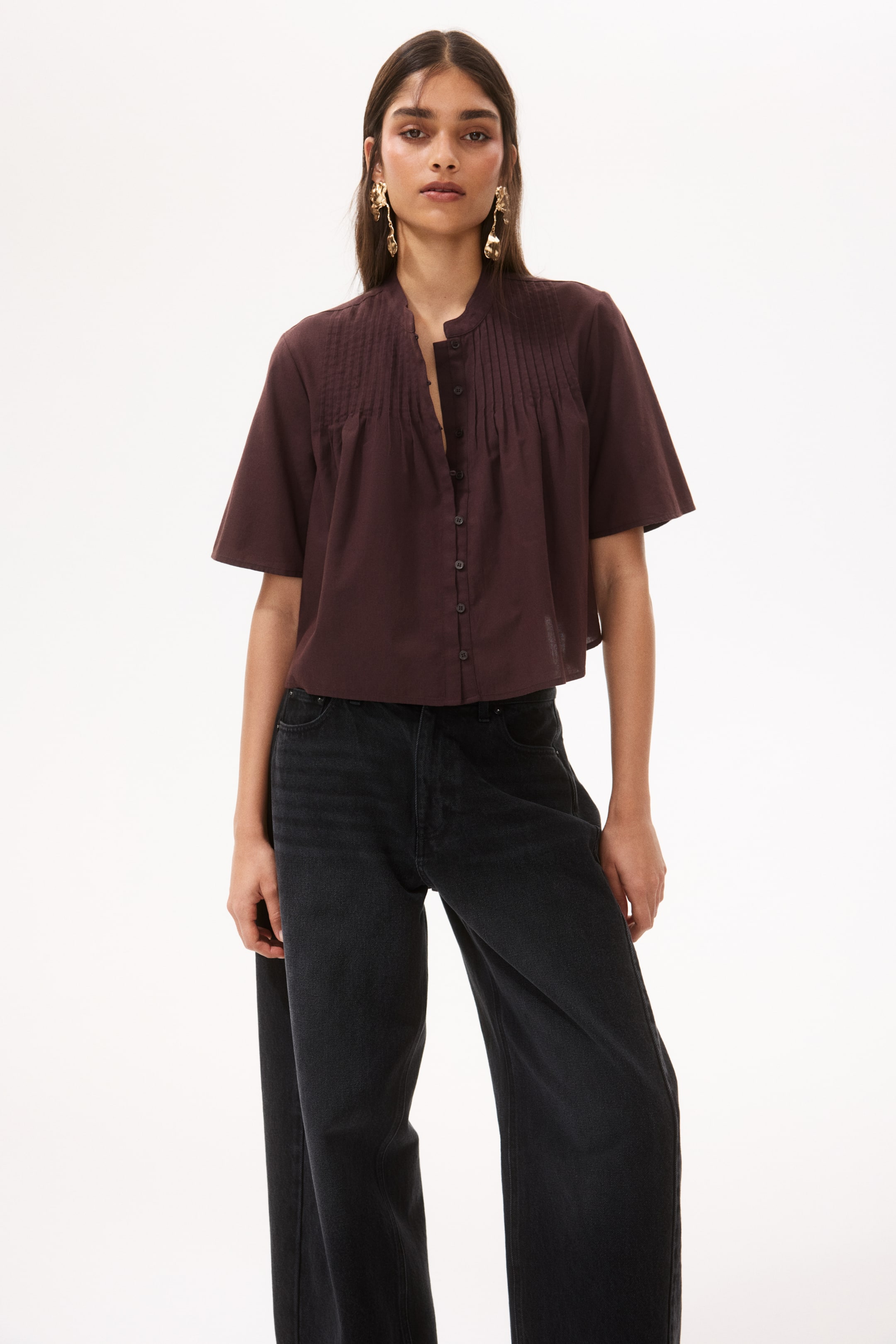 View larger image: A dark brown short-sleeved top featuring a band collar, pintucks on the front, and a partially unbuttoned placket is paired with black wide-leg jeans.