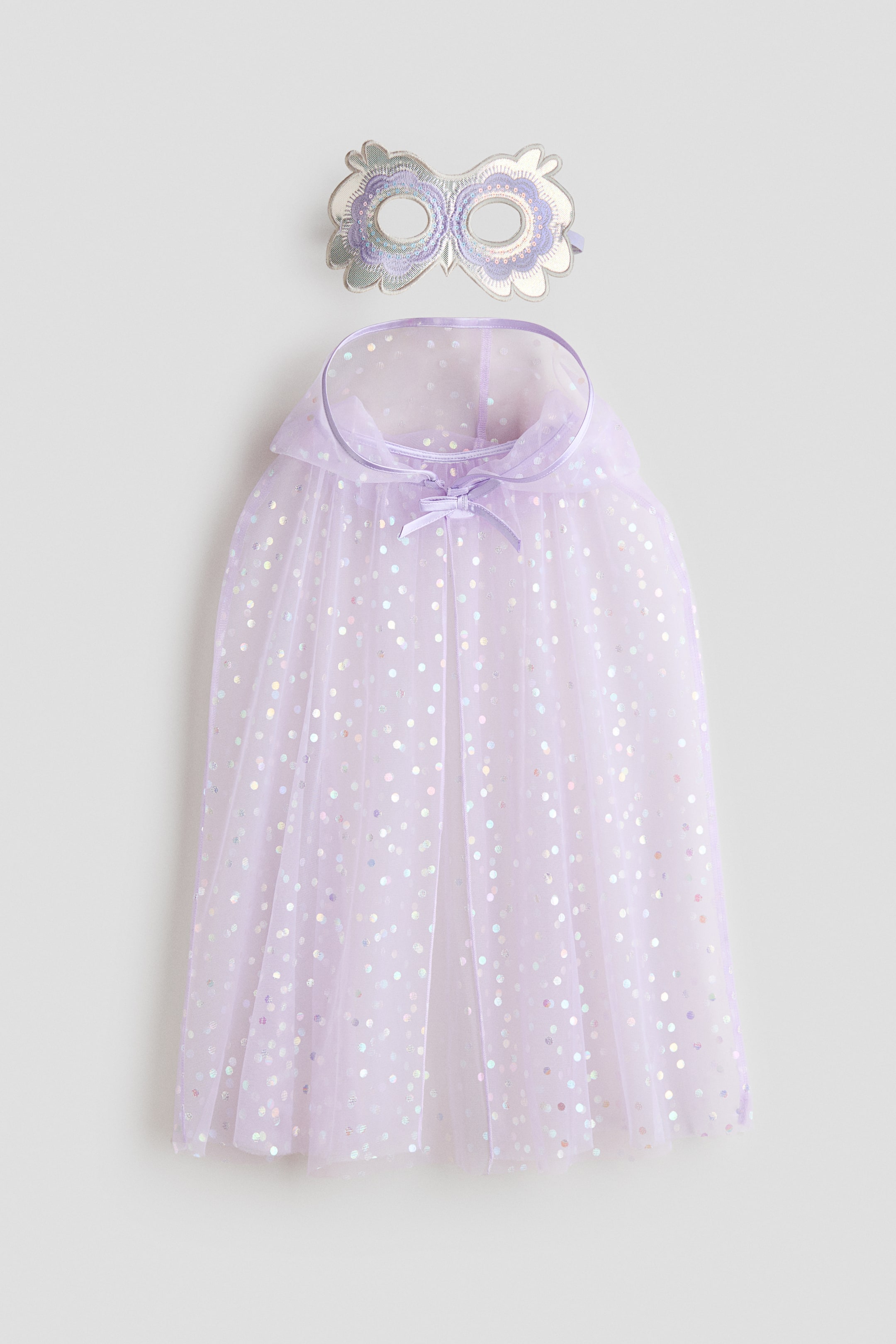 Näytä suurempi kuva: Tuote on etupuolelle suunnattu laventeliasu, joka koostuu läpikuultavasta viitasta ja yhteensopivasta naamiosta. Viitta on koristeltu hohtavilla pyöreillä paljeteilla ja se sidotaan pienellä nauharusetilla kaulan kohdalle, samalla kun naamiossa on pöllömäinen muotoilu hopeisella ja laventelin värisellä kirjailulla.