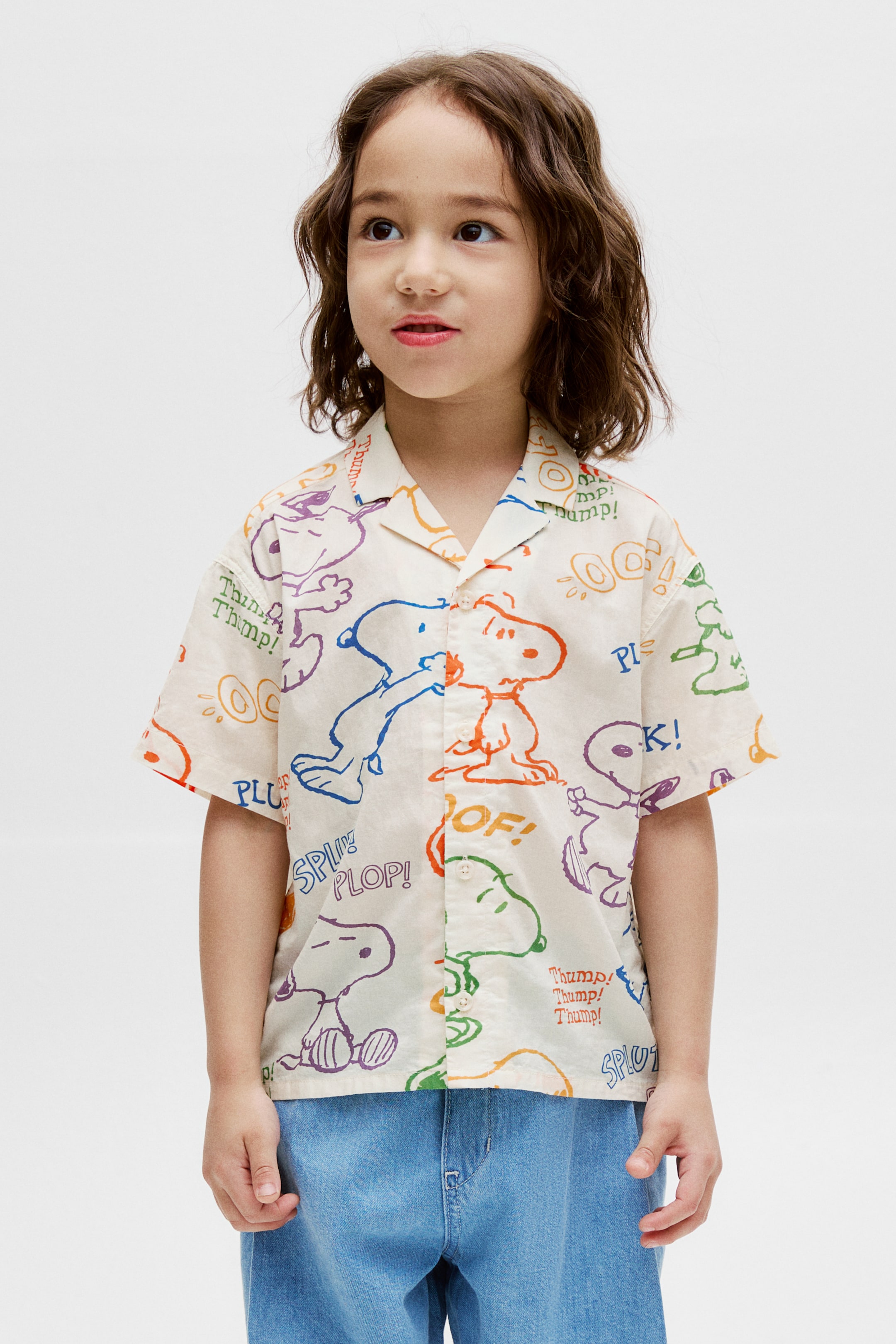 View larger image: A cream-colored short-sleeved shirt is facing frontally, featuring a revere collar and a button-up front. It is adorned with an all-over print of outlined Snoopy and Woodstock characters in various colors, alongside text bubbles with sound effects.
