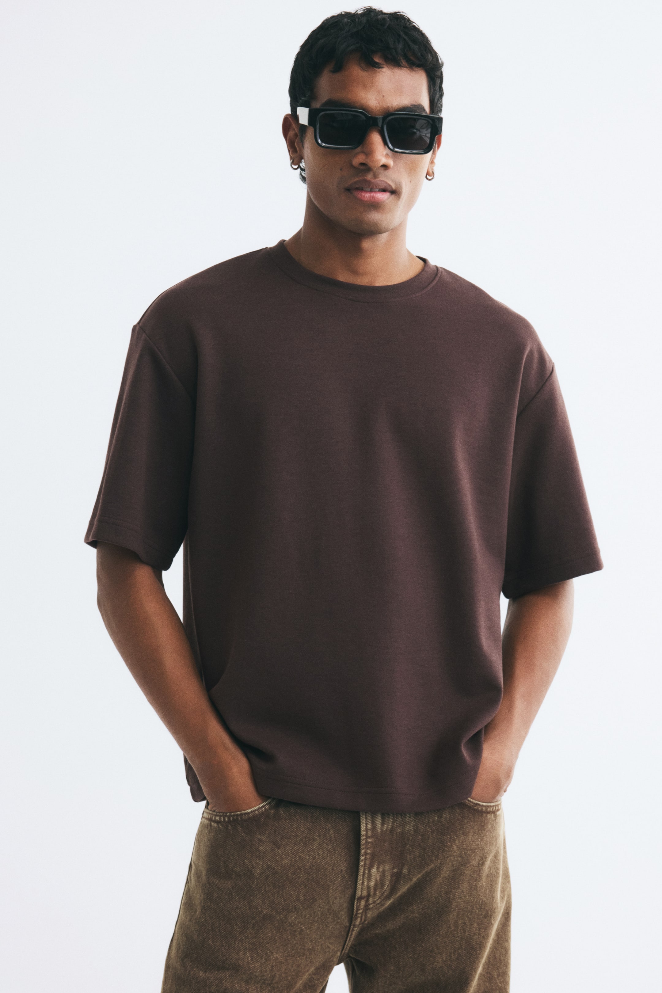View larger image: A dark brown heavyweight jersey T-shirt with a relaxed fit, round rib-trimmed neckline, and dropped shoulders is worn by the model. The model is standing with hands placed in the pockets of brown denim jeans.