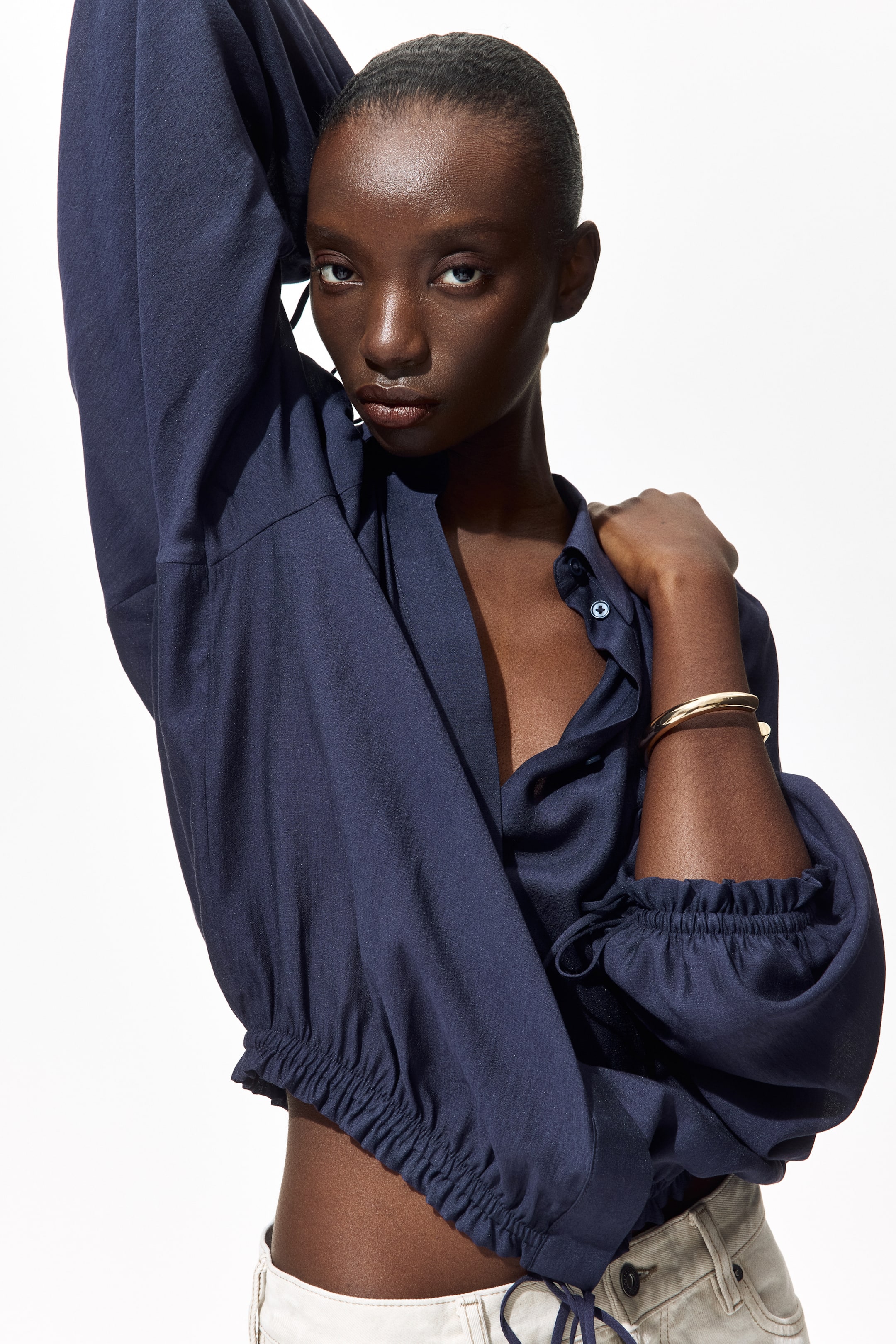 View larger image: The dark blue long-sleeve top, crafted from a textured fabric, showcases an open collared neckline and a partially unbuttoned front. It features voluminous sleeves, gathered cuffs, and an elasticated cropped hem with visible drawstrings.