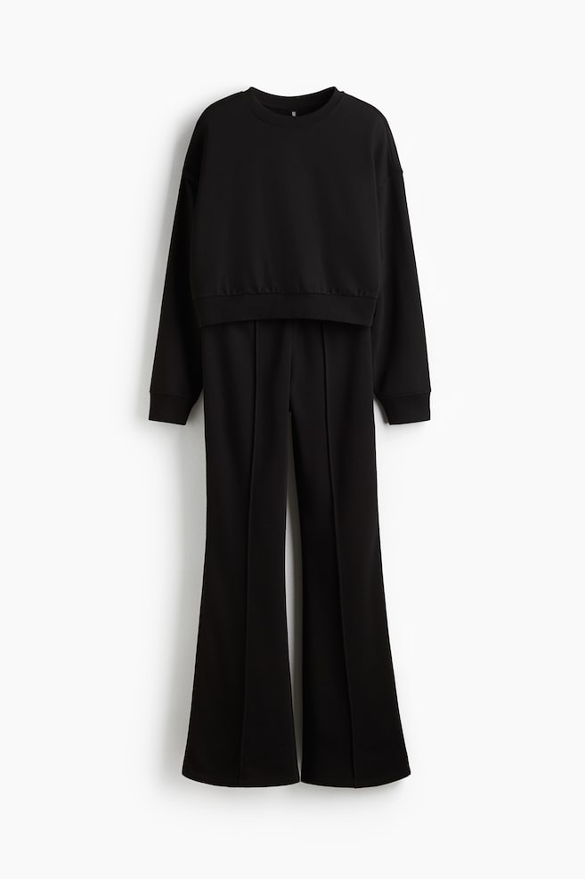 A black two-piece set is presented from the front, featuring a long-sleeved, crew-neck sweatshirt with a ribbed hem and cuffs, along with matching flared trousers that are showing a distinct front seam on each leg. - 1