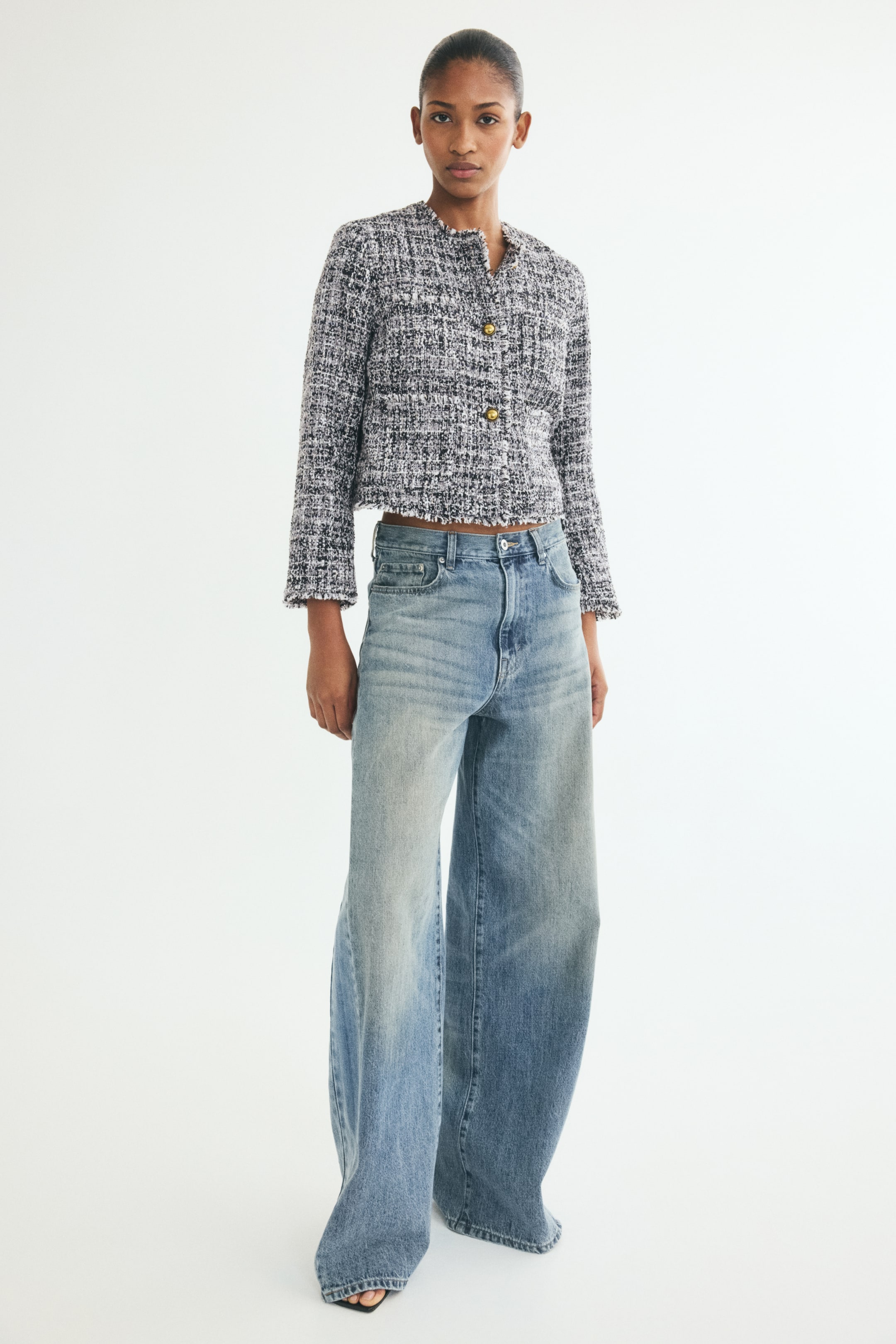 View larger image: A cropped, textured tweed jacket in black and white features gold-tone button closures and a frayed hem, paired with high-waisted, wide-leg jeans in a light blue wash. The front-facing view showcases the structured fit of the jacket and the relaxed drape of the denim.