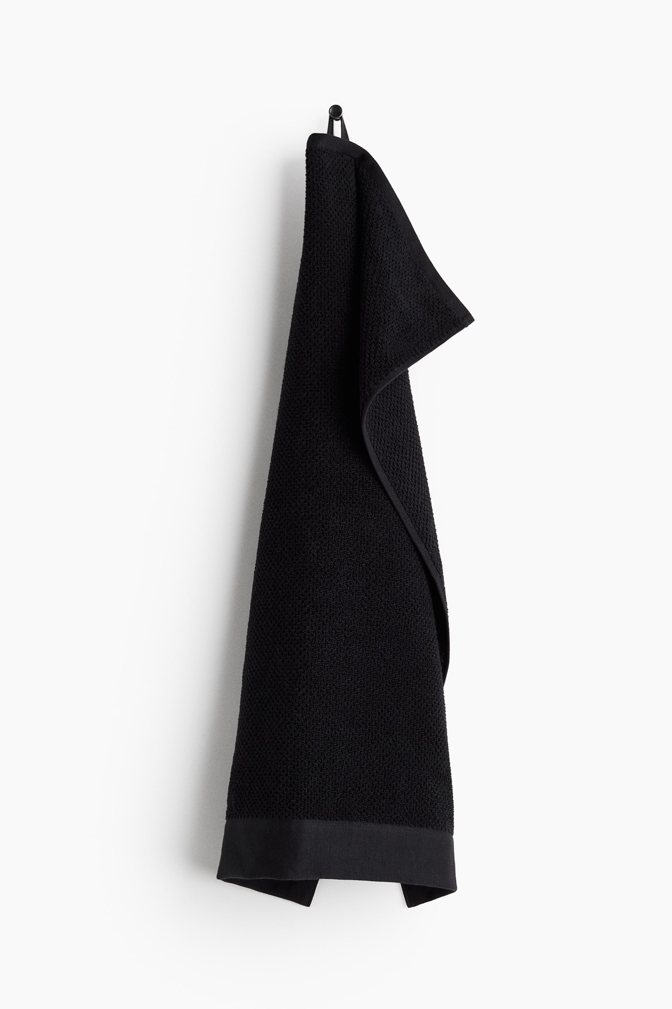 View larger image: A black hand towel is hanging vertically from a hook, showcasing a textured terry weave. It has a finished band along its bottom edge, and a loop is visibly attached at the top for easy hanging.