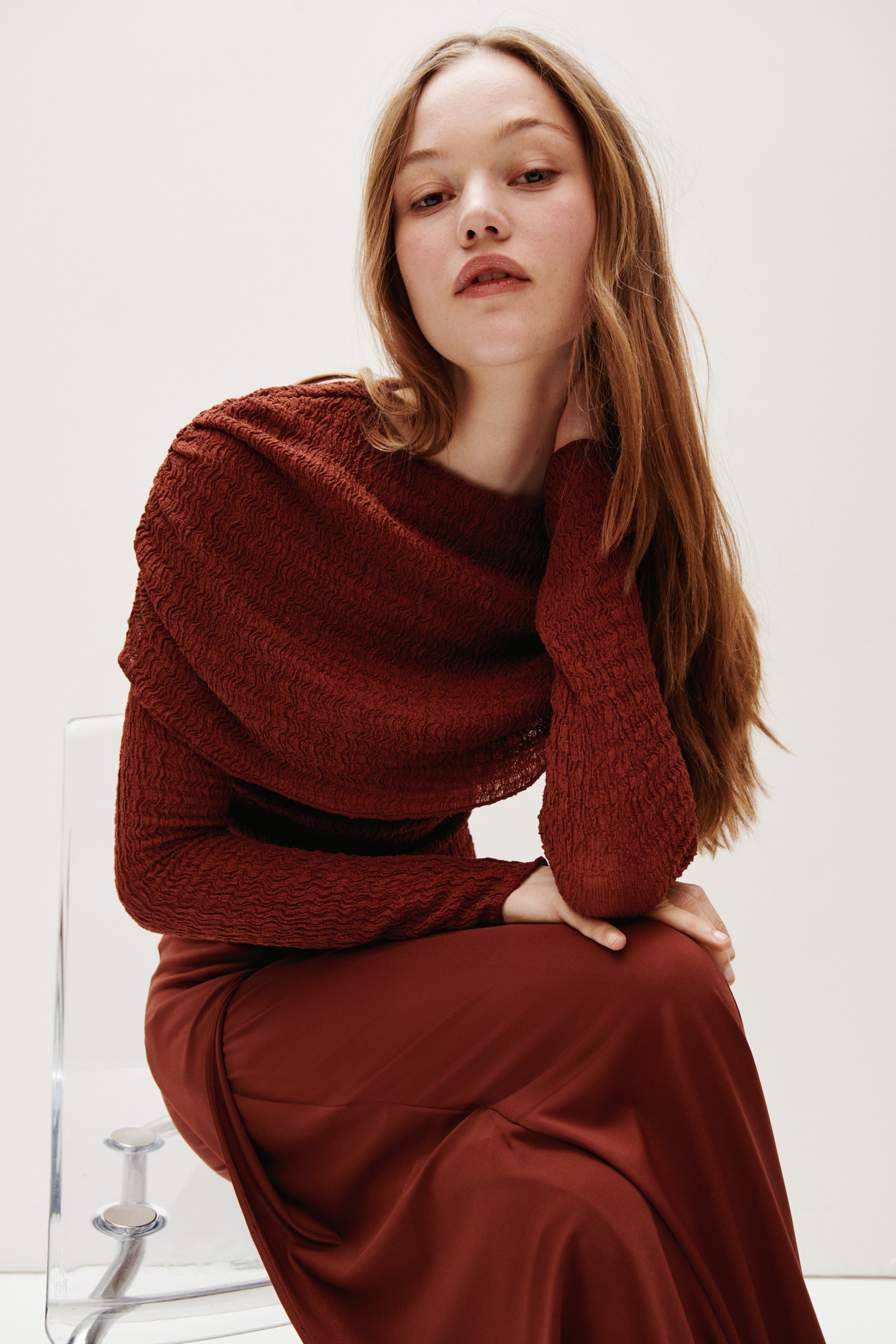 Ingrandisci l'immagine: Un top in tessuto arricciato di un marrone rossastro scuro presenta uno scollo drappeggiato e asimmetrico e maniche lunghe. È abbinato a una gonna liscia e fluida dello stesso colore, che mostra un sottile dettaglio drappeggiato sul fianco sinistro.