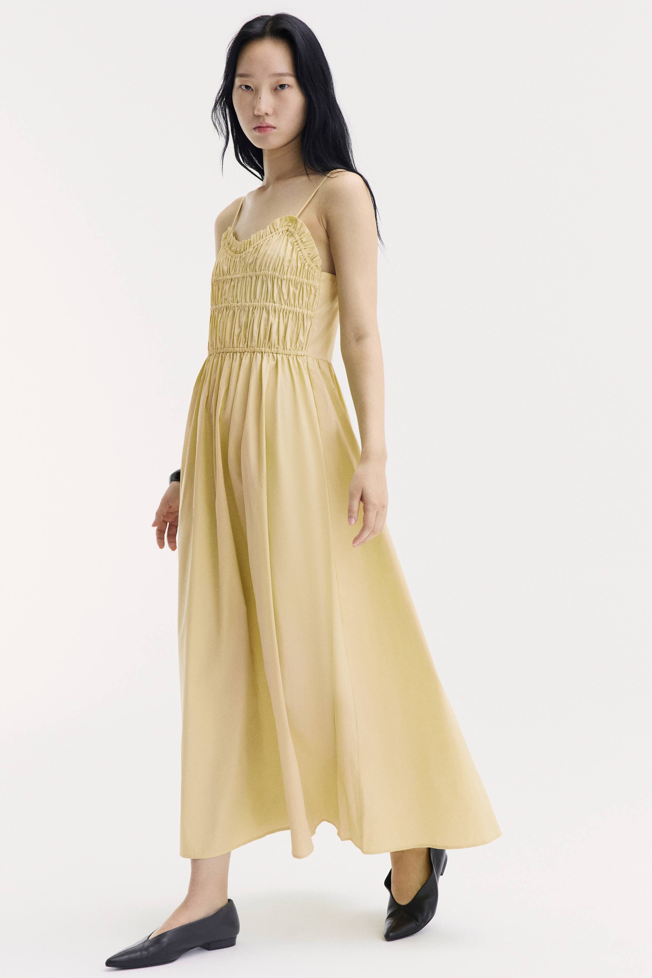 View larger image: A light yellow, calf-length dress is worn by the model, featuring a ruched bodice with horizontal elasticated seams, thin shoulder straps, and a flared, flowing skirt. The model is standing and posing in the dress, which is styled with black pointed-toe flat shoes.
