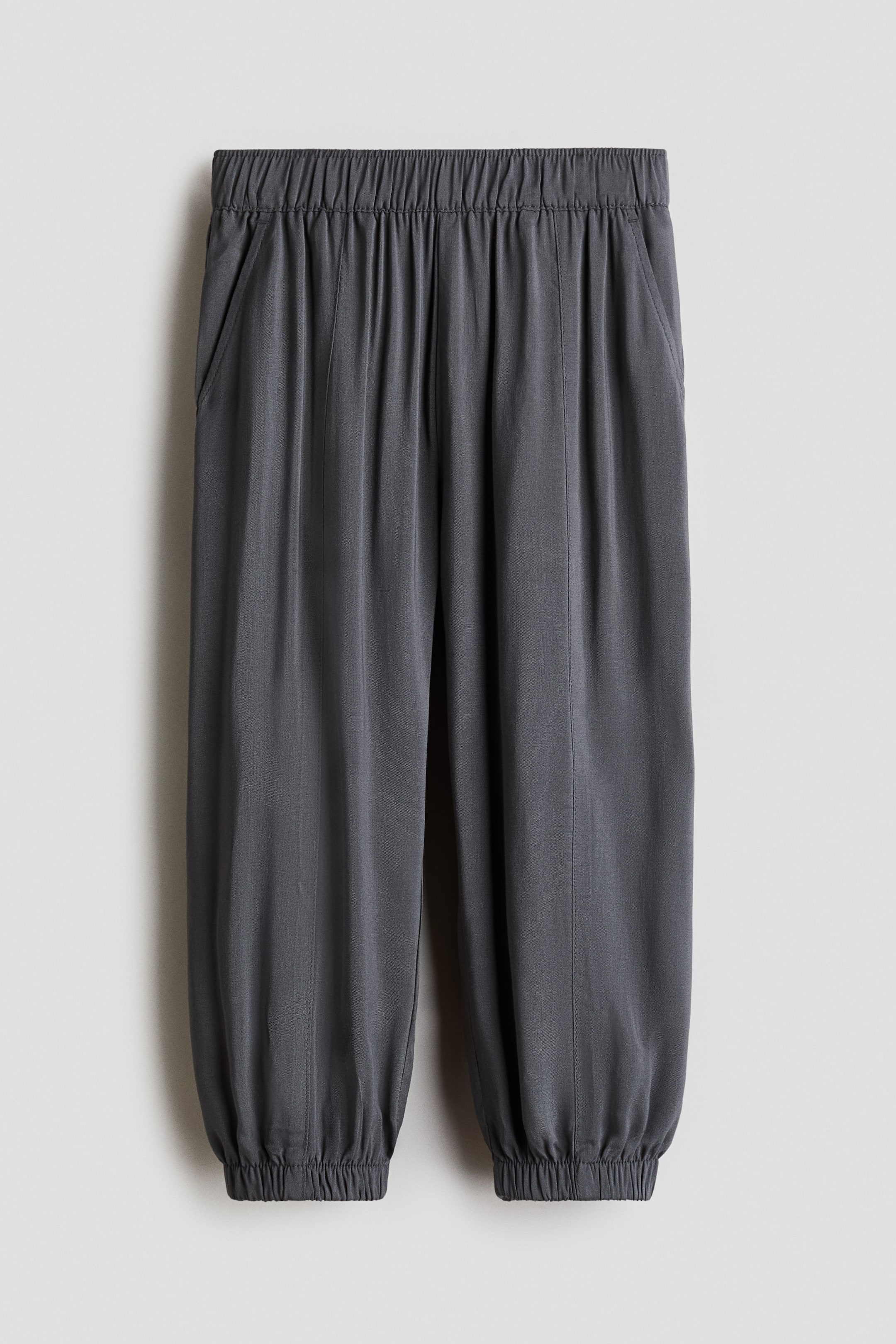 拡大画像を表示: バルーンパンツは、濃いグレーのビスコース織りで作られたフロントからのデザインです。ウエストはゴム仕様で、斜めのサイドポケットがあり、裾にもゴムが施されています。