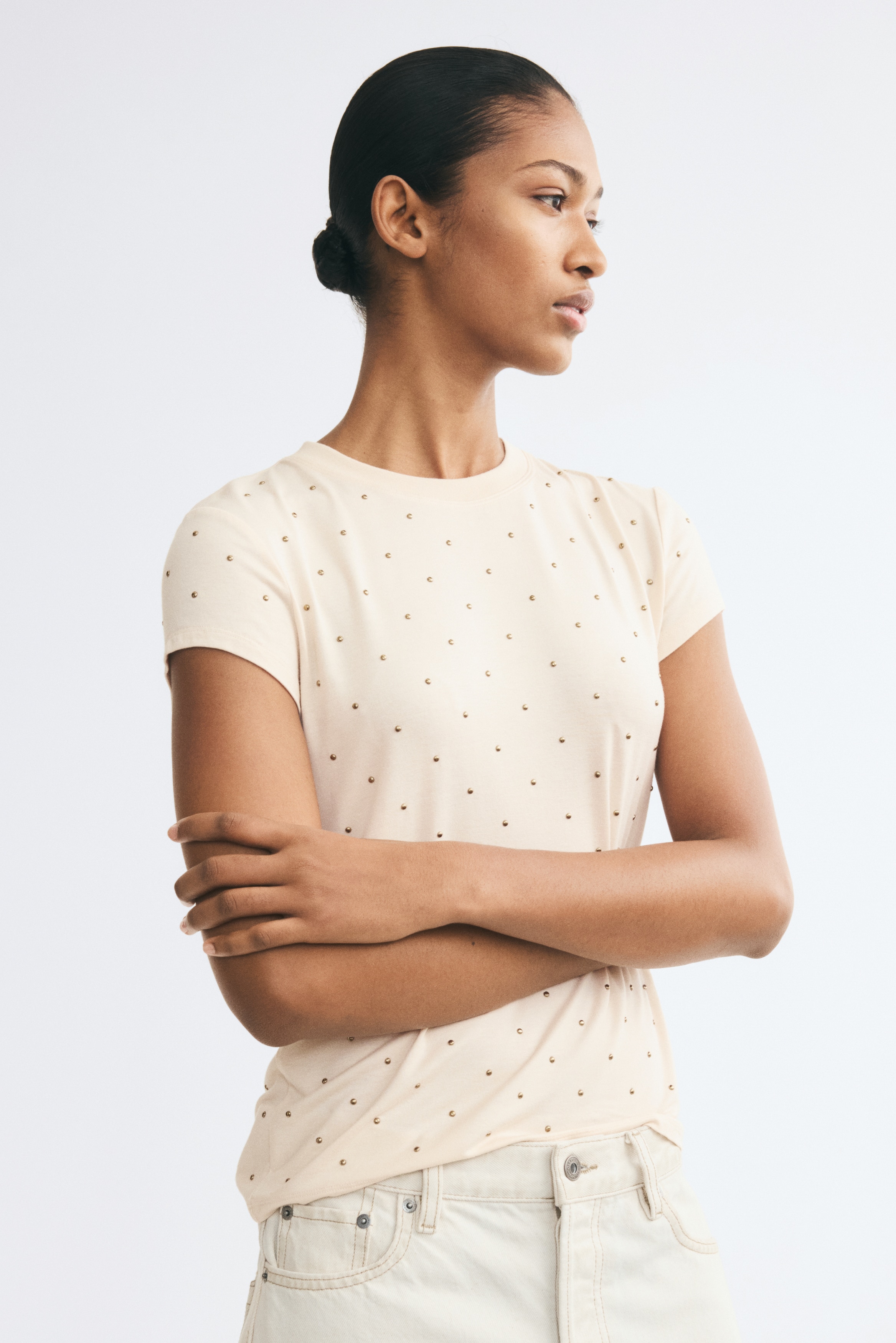 A cream-coloured, short-sleeved T-shirt is shown from the front, featuring a classic crew neckline and a straight hem. Small, evenly spaced gold metallic studs embellish the entire front of the garment and its sleeves.