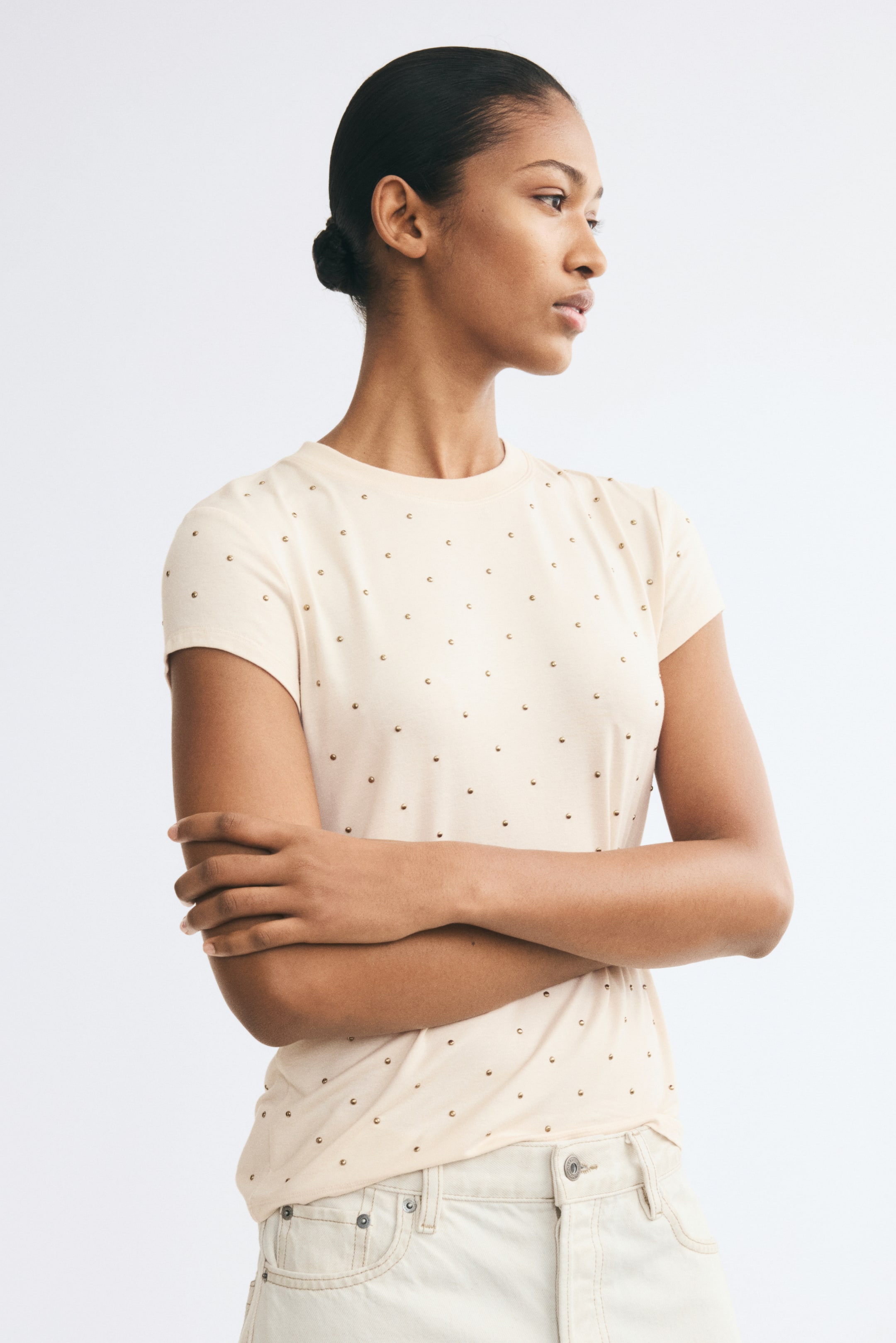 View larger image: The model is wearing a light beige fitted t-shirt adorned with small, shiny metallic studs, posing with her arms crossed. The tee features a round, rib-trimmed neckline and short sleeves, and is styled with light-colored denim pants.