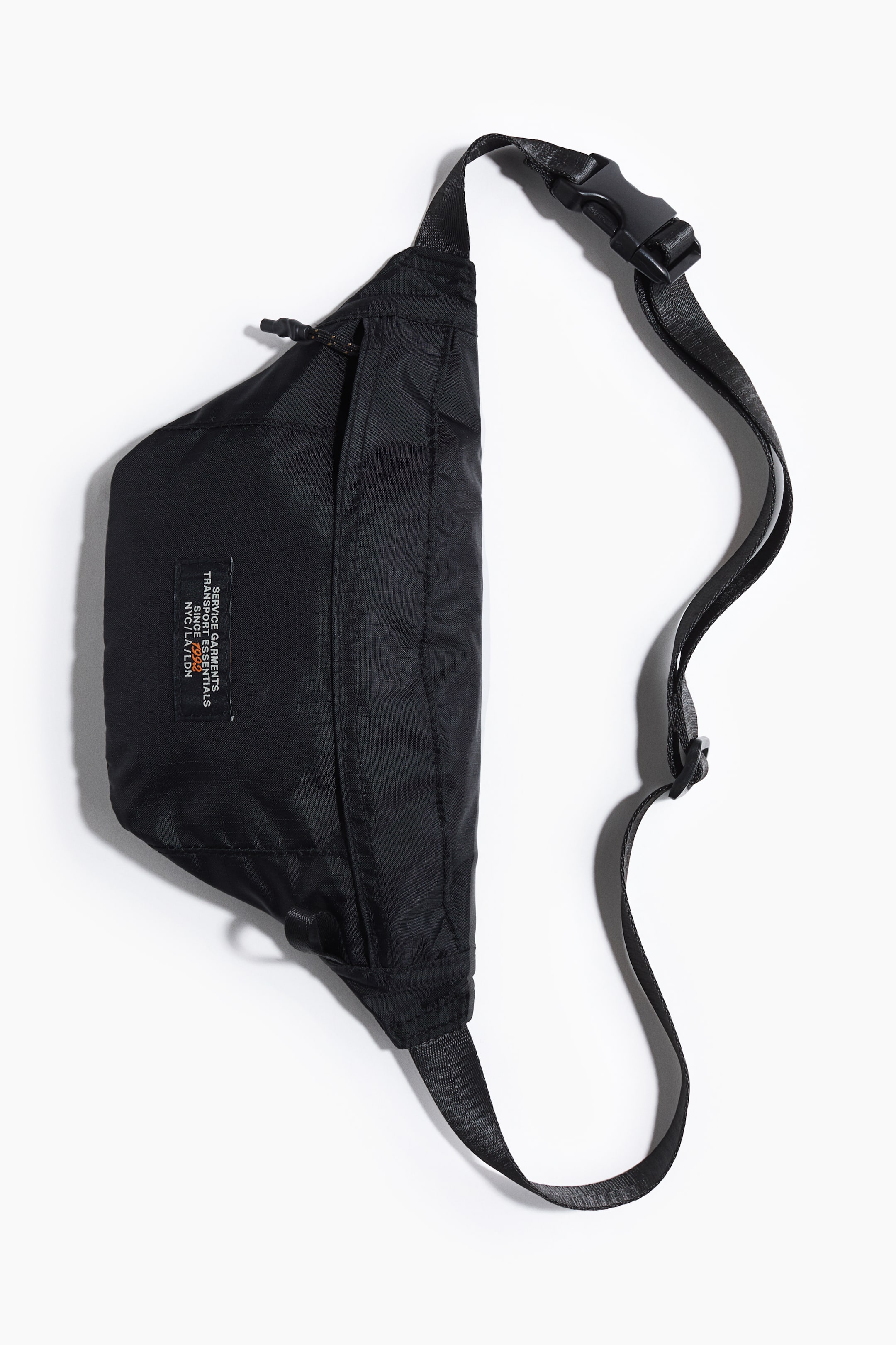 Ver imagen más grande: Una riñonera negra se presenta de frente, con un compartimento principal con cremallera, un bolsillo delantero más pequeño con cremallera y un tirador de cordón, y una etiqueta rectangular blanca con texto naranja. Una correa ajustable de cinta negra con una hebilla de plástico se extiende desde el bolso.