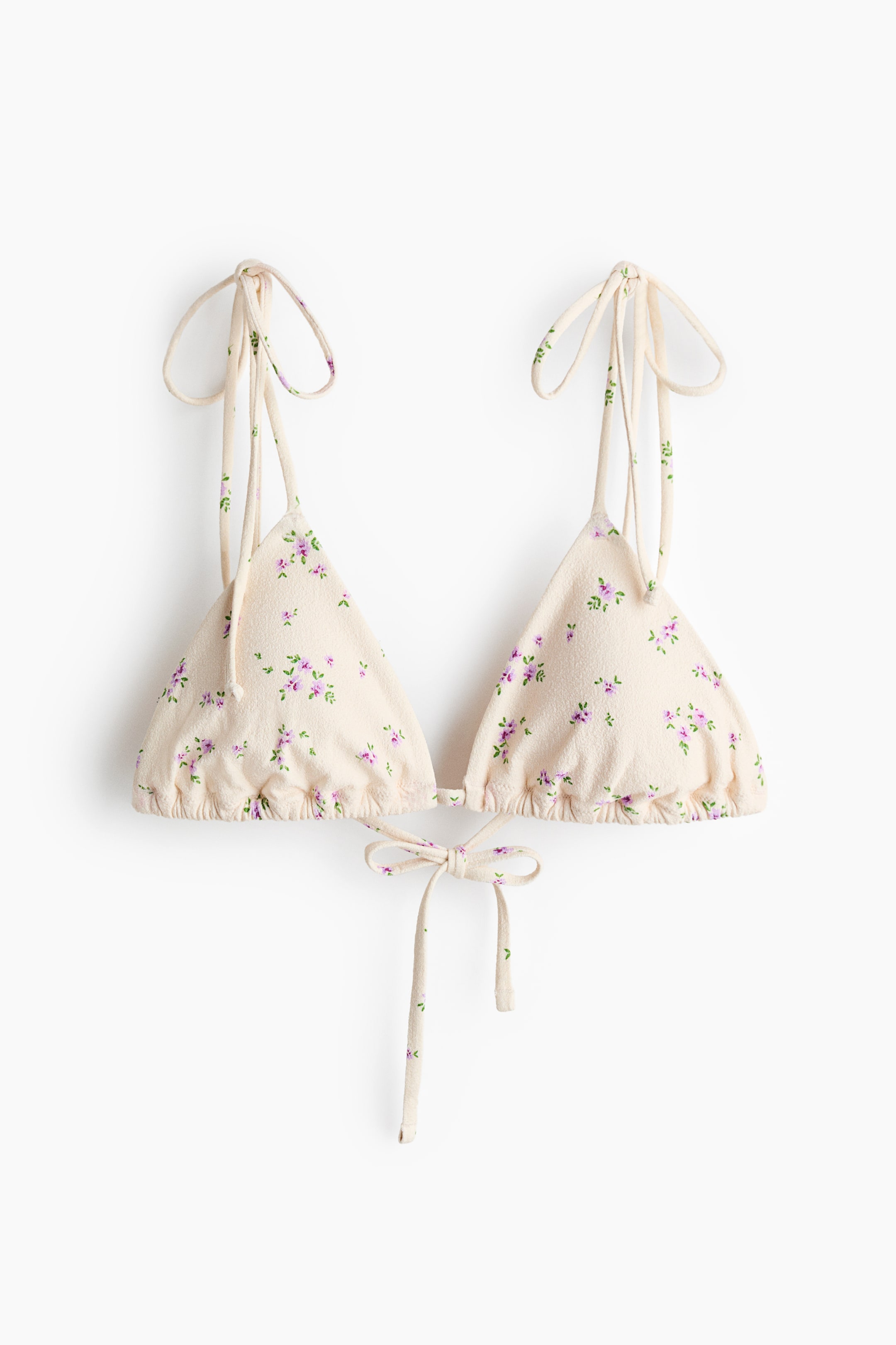 Se større billede: Et trekantet bikini-top med frontvendt design fremviser en cremefarvet stof med små lilla og grønne blomsterprint. Den har bindebånds skulderstropper og en bagpå-lukning, der danner en sløjfe i midten.