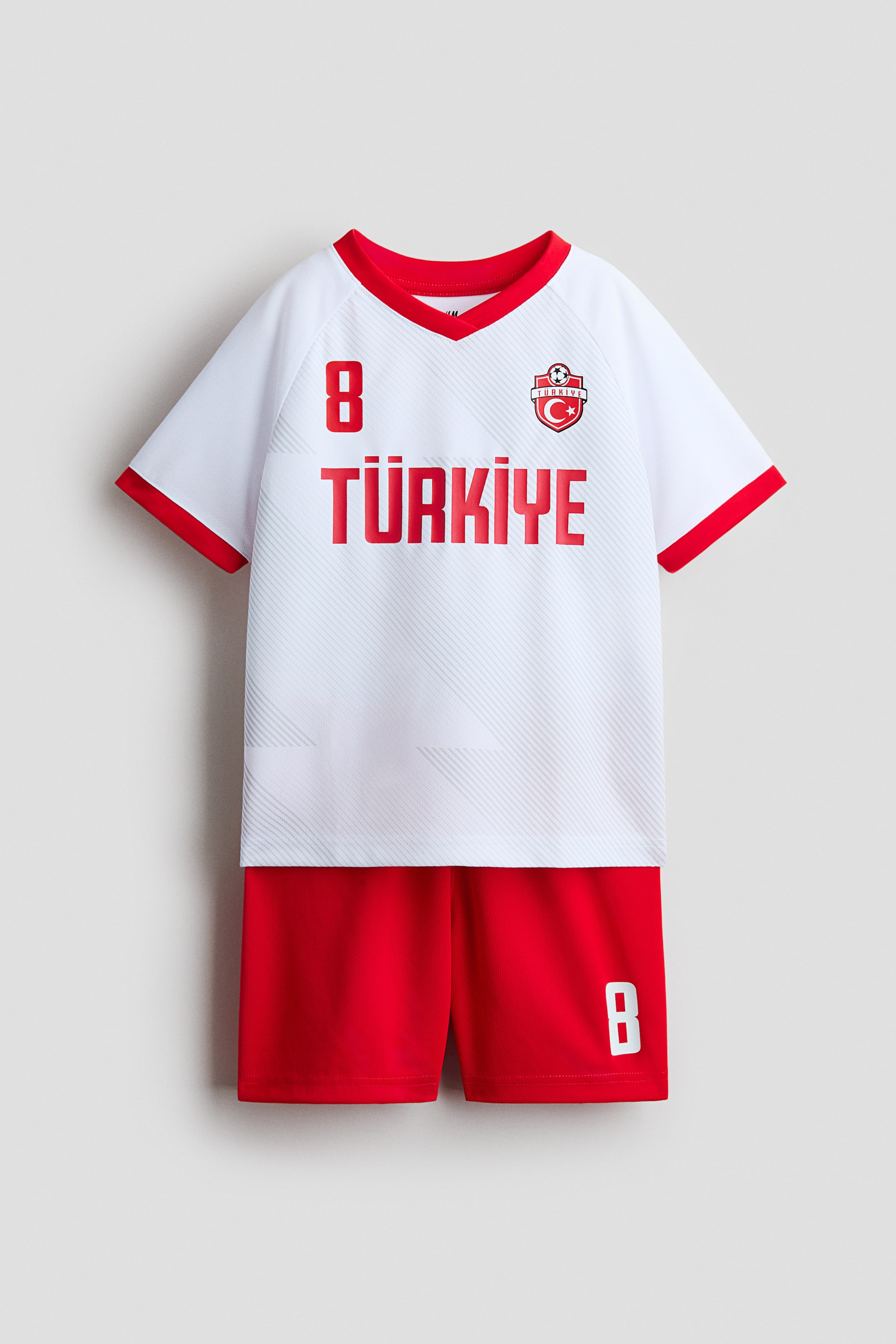 Büyük resmi görüntüle: Yumuşak file kumaştan üretilmiş baskılı bir futbol seti, ön taraftan gösterilmektedir. Beyaz kısa kollu üst, V yaka ve kol manşetlerinde kırmızı şeritlerle tasarlanmıştır. Üst kısımda kırmızı 'TÜRKİYE' yazısı ve göğüste kırmızı '8' numarası bulunmaktadır. Eşlik eden kırmızı şortlar ise sağ bacakta beyaz '8' numarası ve lastikli, bağcıklı bir bel kısmı ile tasarlanmıştır.