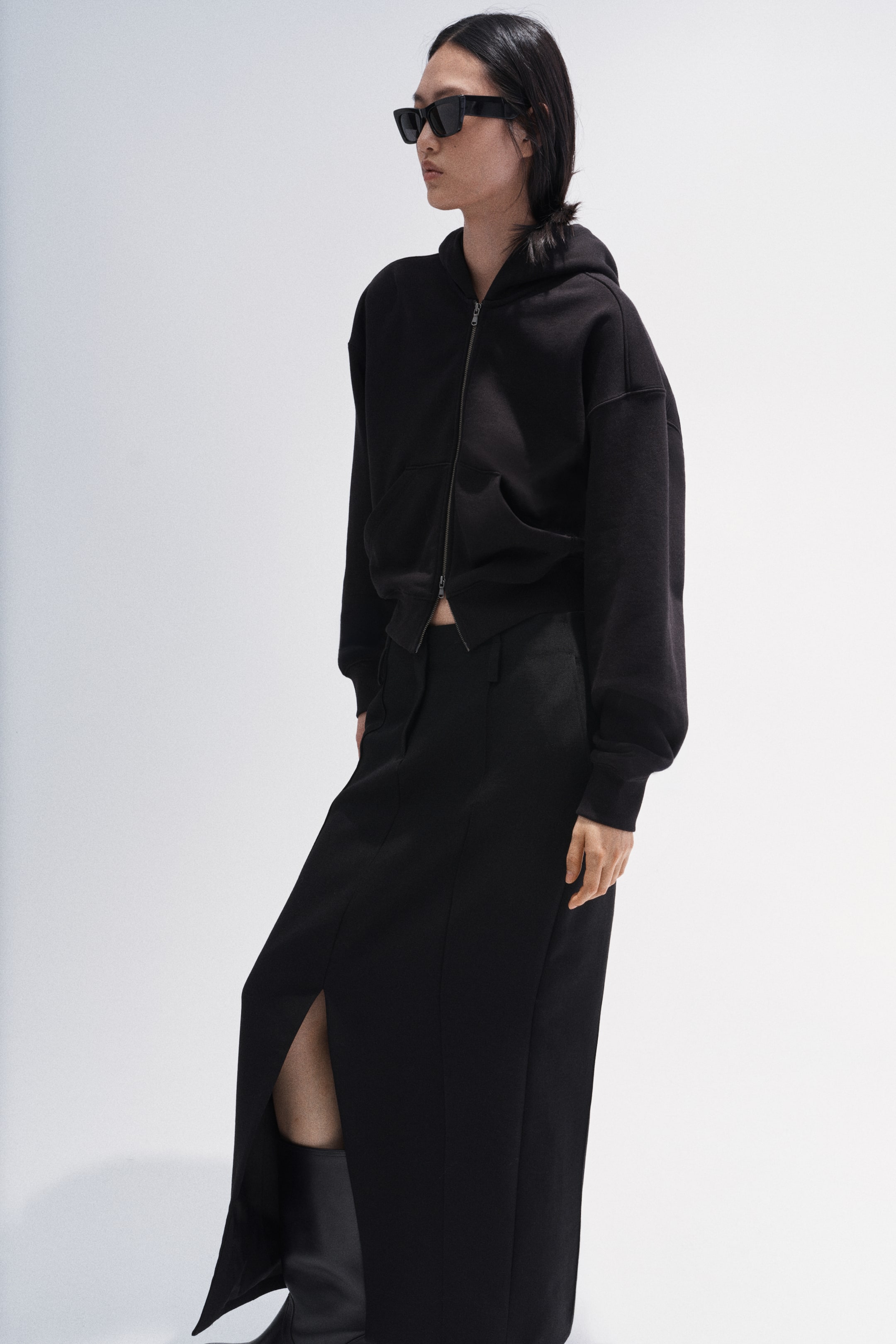 View larger image: A black cropped zip-up hoodie is being worn by the model, featuring a hood, long sleeves, and a front zip closure with two front pockets.