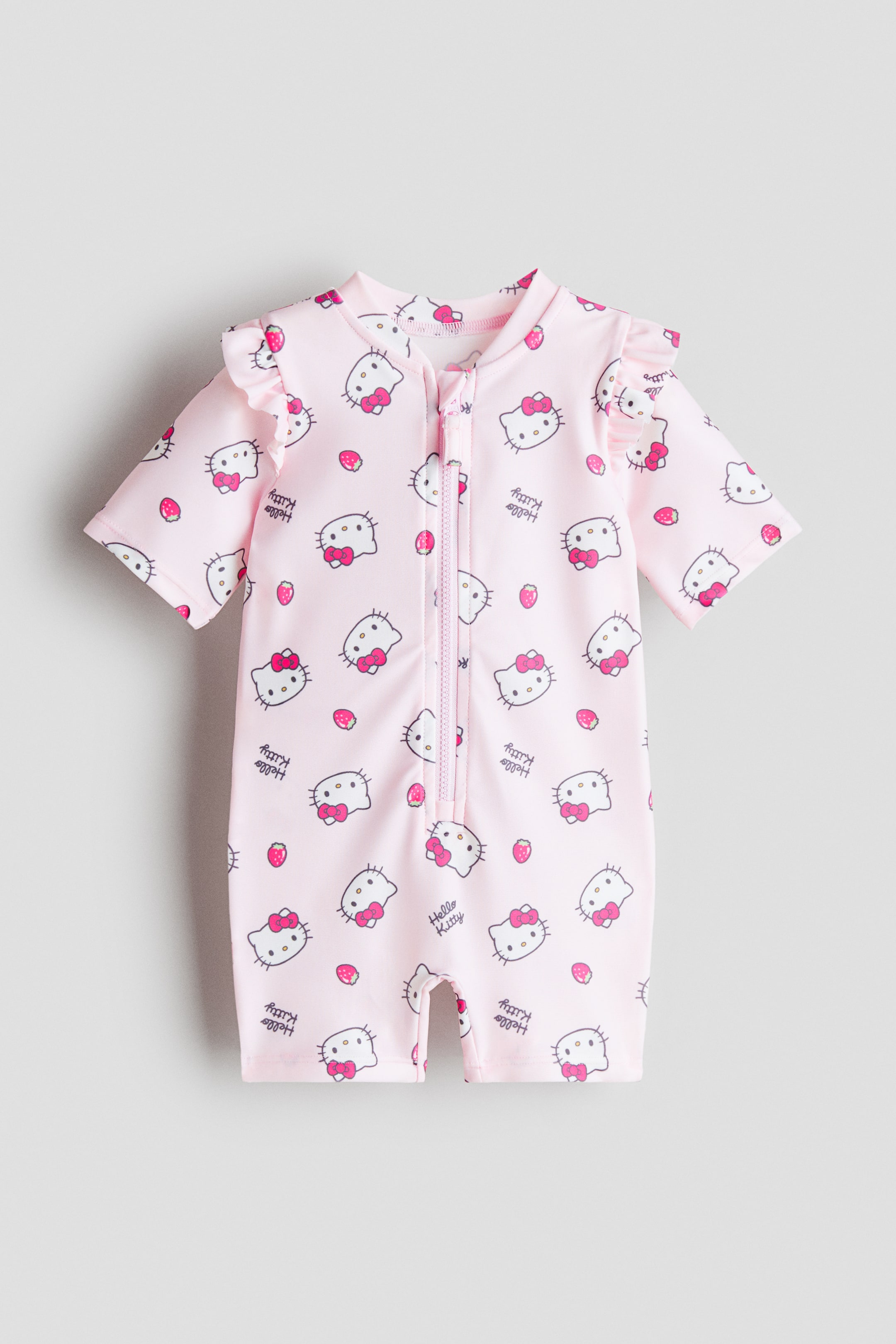 Voir une image plus grande: Un maillot de bain une-pièce rose est présenté de face, avec un imprimé intégral de visages Hello Kitty, de fraises rouges et du texte « Hello Kitty ». Il est conçu avec des manches courtes à volants, des jambes courtes, un col rond montant et une fermeture éclair frontale sur toute la longueur.