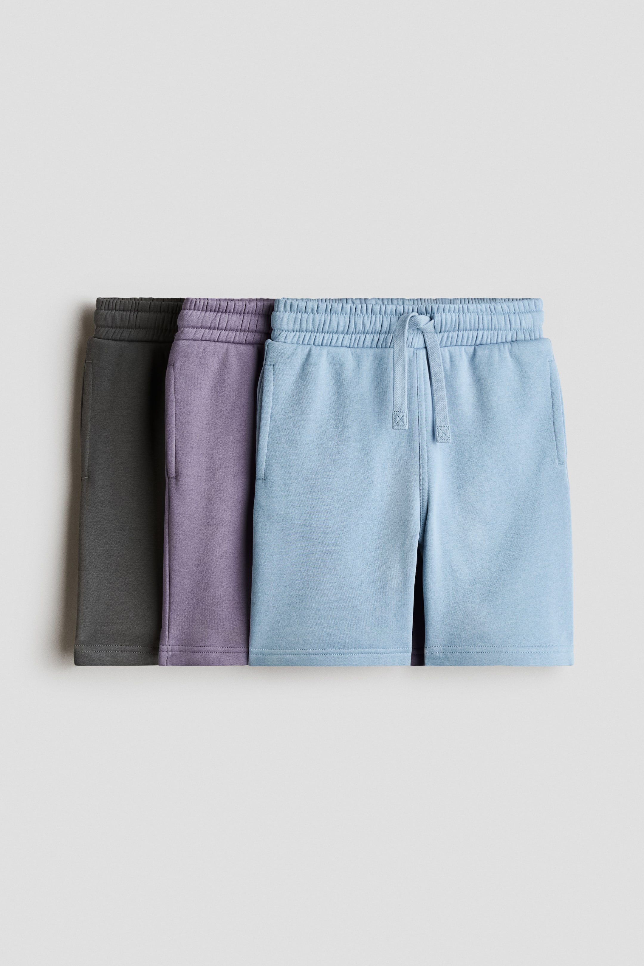 Grotere afbeelding bekijken: Drie paar shorts in lichtgewicht sweatshirtstof worden aan de voorkant getoond. Elk paar heeft een elastische tailleband met trekkoord en discrete zijzakken, en is verkrijgbaar in donkergrijs, lichtpaars en lichtblauw.