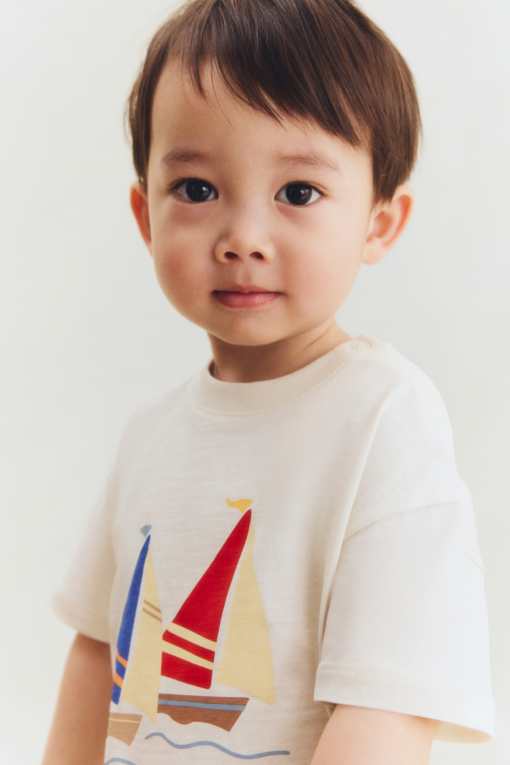 Agrandir l'image: Un t-shirt crème clair présente un petit motif brodé de trois voiliers colorés sur le devant. Il est conçu avec un col rond à bord-côte avec des boutons-pression sur une épaule, et des épaules tombantes.