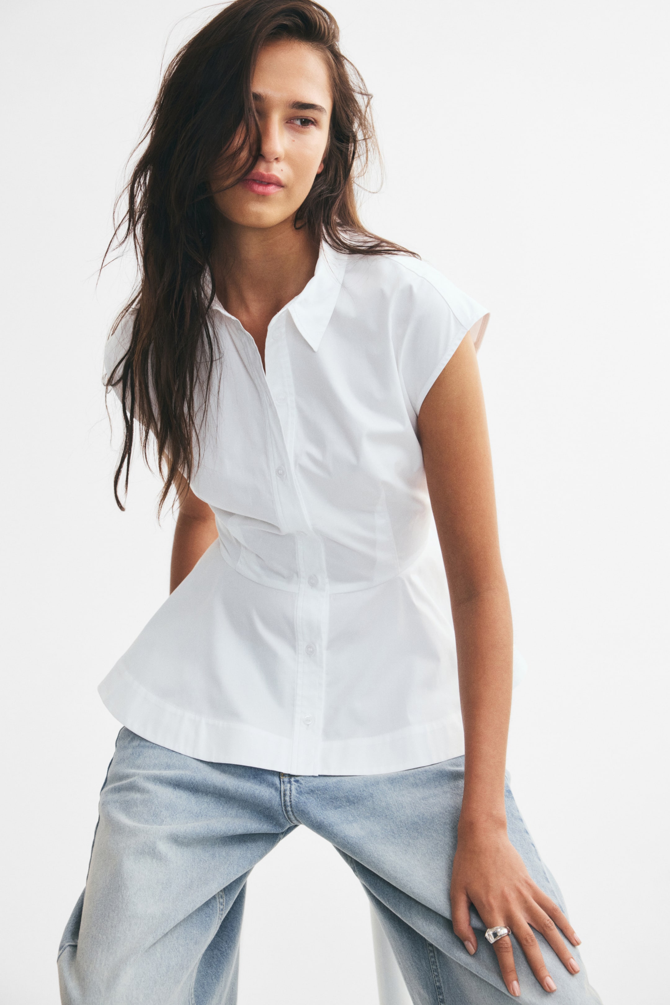 View larger image: A sleeveless white cotton poplin top with a collar, front button closures, and a flared peplum hem is worn by the model. The top is fitted at the waist with vertical darts and paired with light-wash denim jeans.