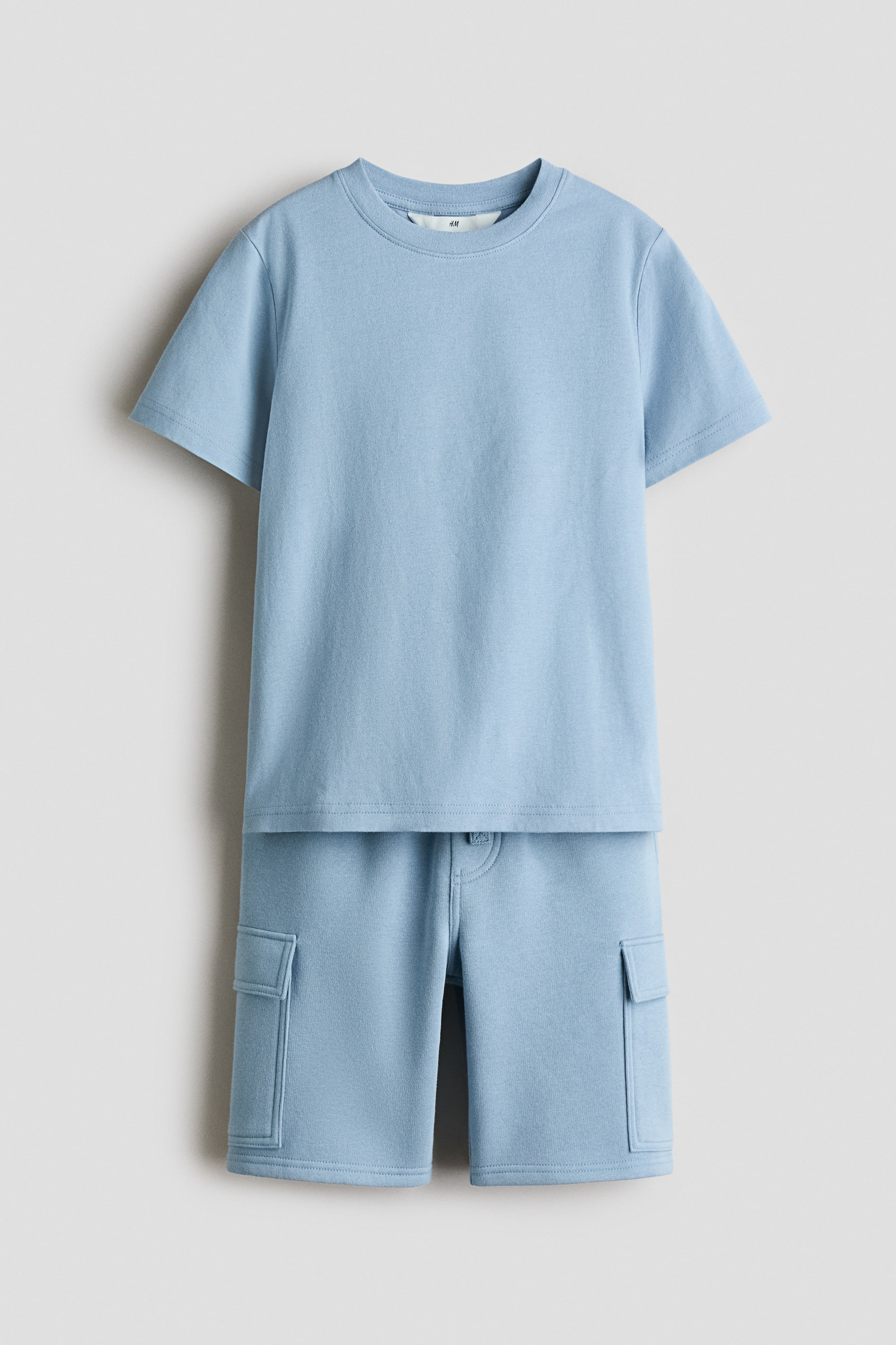 Ver imagen más grande: Conjunto de camiseta y pantalones cortos en azul claro mostrando una vista frontal. La camiseta está confeccionada en suave algodón jersey, mientras que los pantalones cortos están hechos de tejido de sudadera. La camiseta presenta un cuello redondo con ribete y un dobladillo recto, mientras que los pantalones cortos cuentan con una cintura elástica con cordón, una bragueta falsa, bolsillos laterales diagonales y bolsillos de parche con solapas en las piernas.