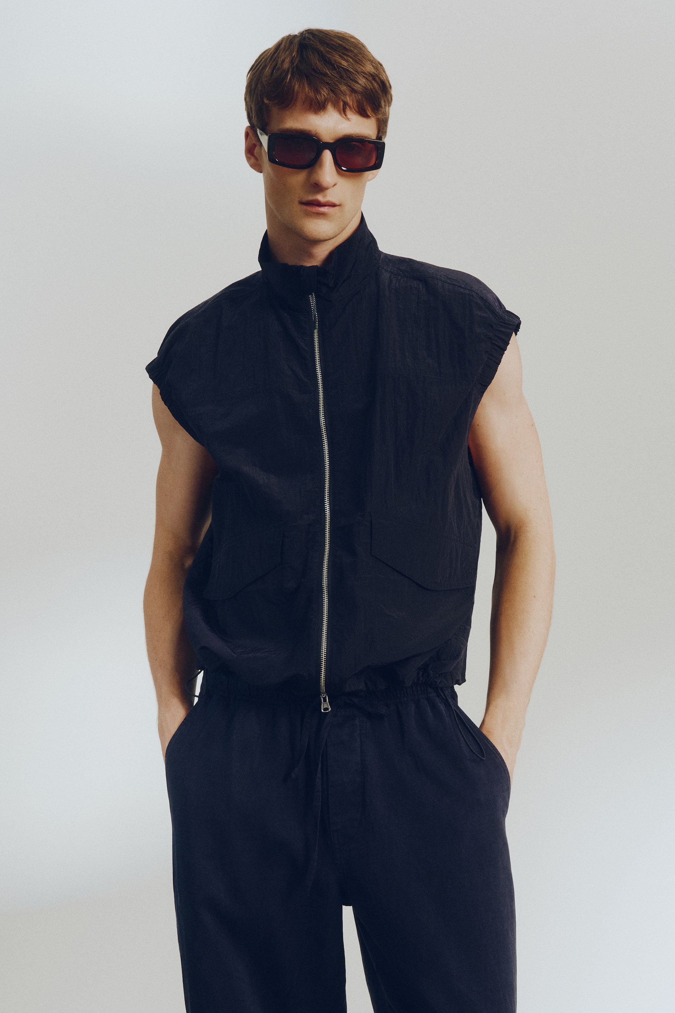 Agrandir l'image: Un gilet sans manches froissé de couleur sombre est présenté, doté d'un col montant, d'une fermeture éclair argentée à l'avant, de deux poches à rabat et d'un ourlet élastiqué. Un pantalon assorti froissé complète l'ensemble, avec une taille à cordon de serrage et des poches latérales.