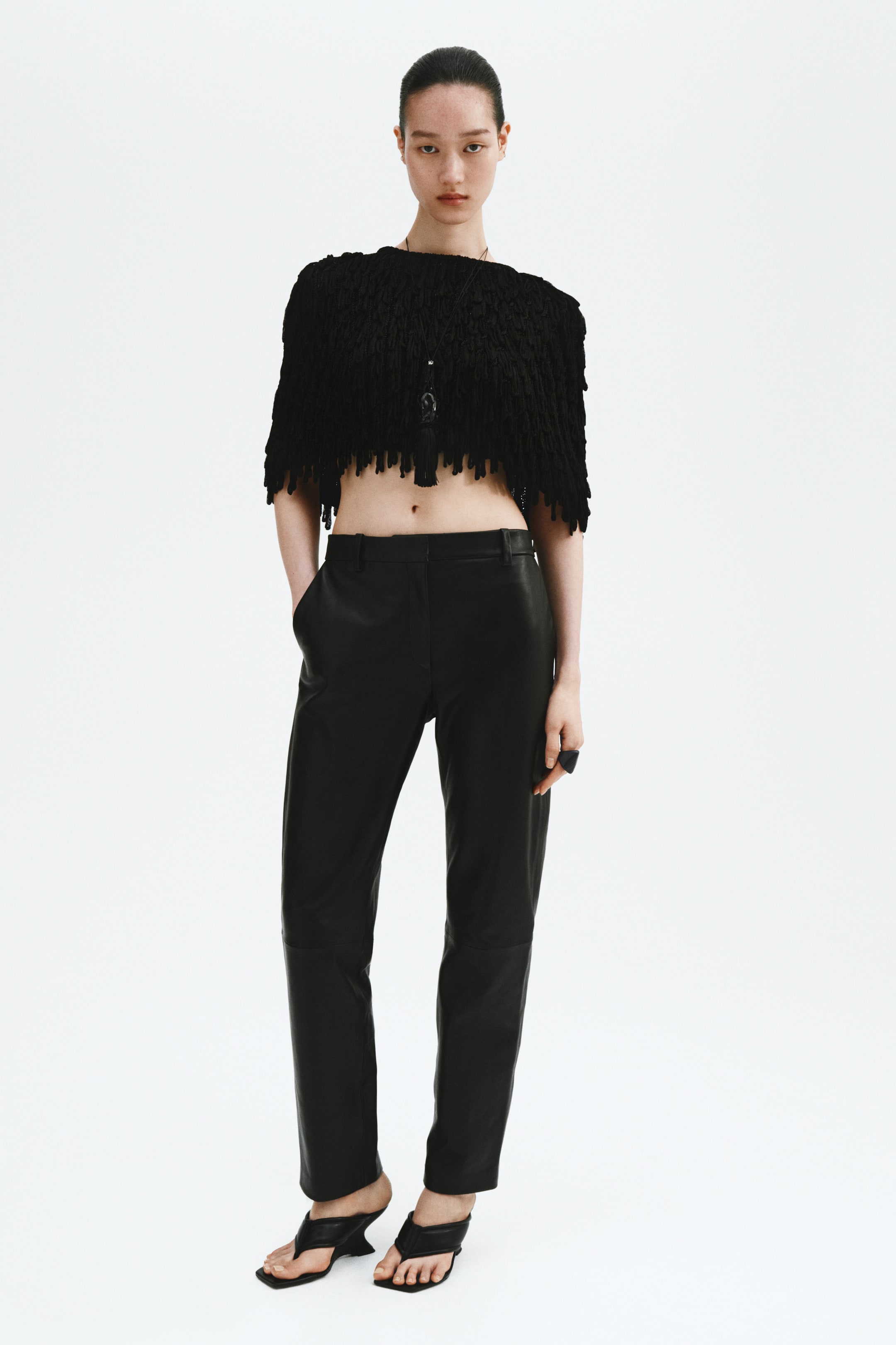 View larger image: A black textured cropped top features a dense, shaggy surface and an irregular fringed hemline, with a necklace and black tassel pendant resting on the front. Black leather pants showcase a straight-leg fit and seam details at the knees, with hands placed in the side pockets.