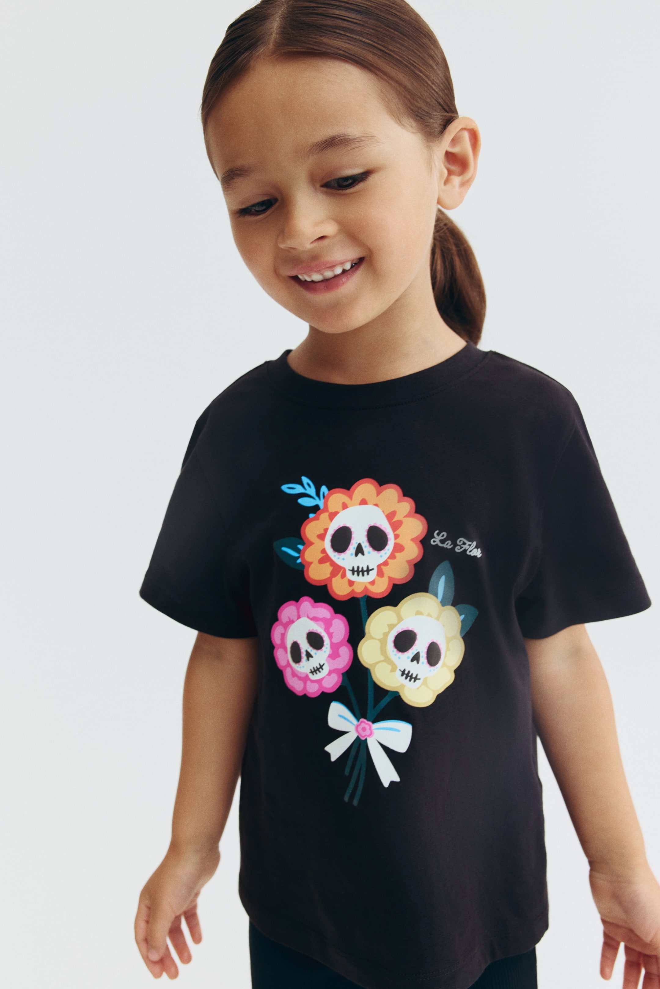 View larger image: A black cotton jersey tee is being worn facing forward, featuring a crew neckline and short sleeves. The front of the tee features a colorful graphic print of three skull-headed flowers tied together with a bow.