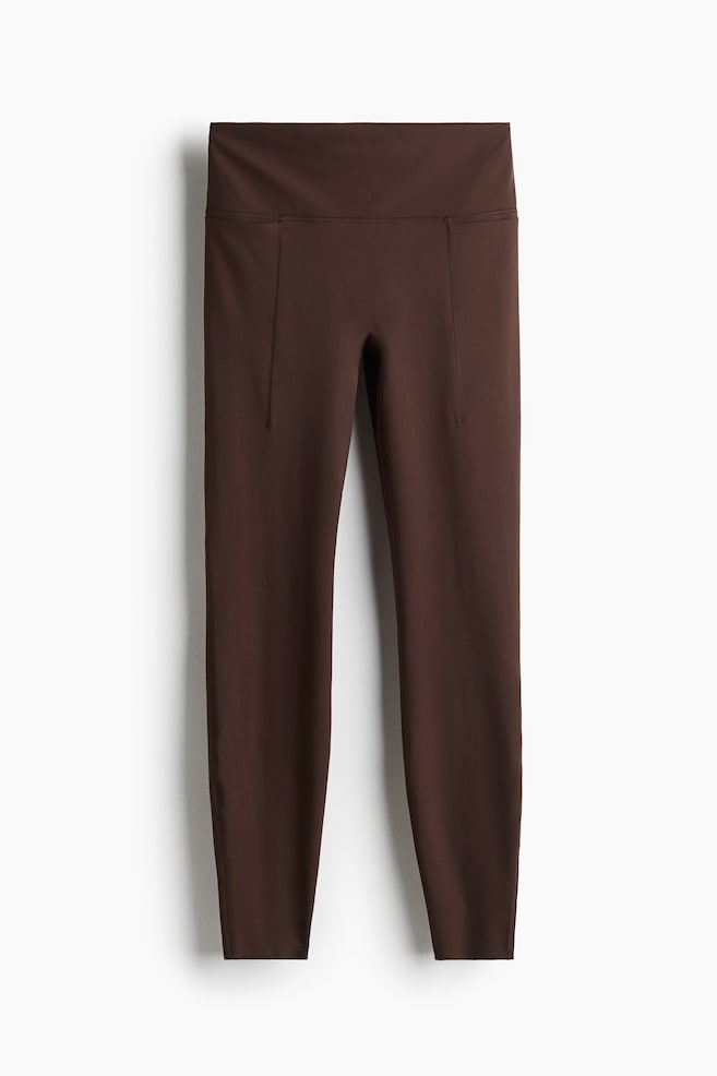 Dark brown leggings are being presented from the front, featuring a wide, high-rise waistband and visible vertical seams running down the front of the legs. - 1
