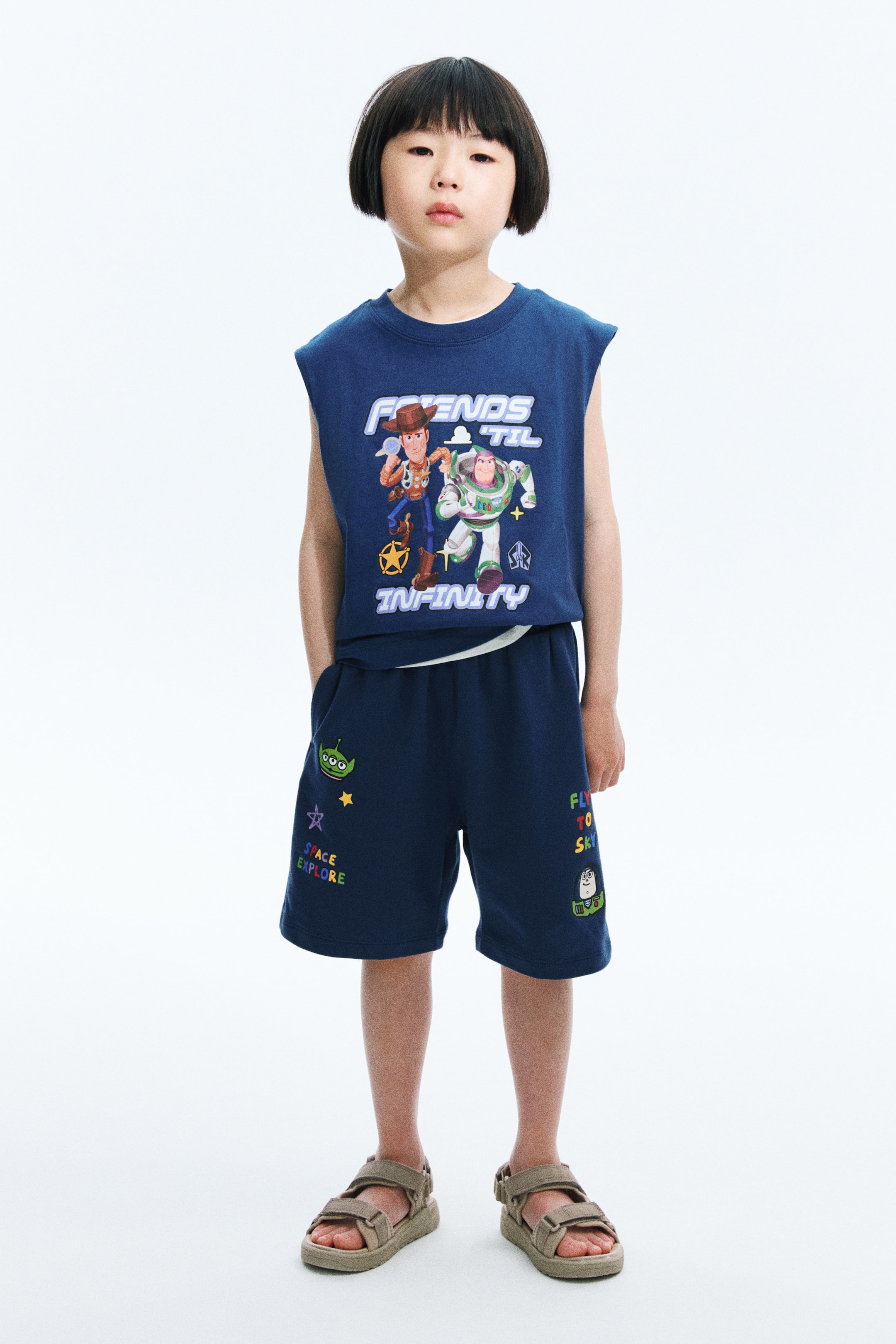 View larger image: A navy blue sleeveless top, showcasing a "Friends 'Til Infinity" graphic with Woody and Buzz Lightyear, paired with matching shorts adorned with vibrant Toy Story-themed graphics like an alien and "Space Explore" text, is being displayed. A white shirt is layered beneath the top.