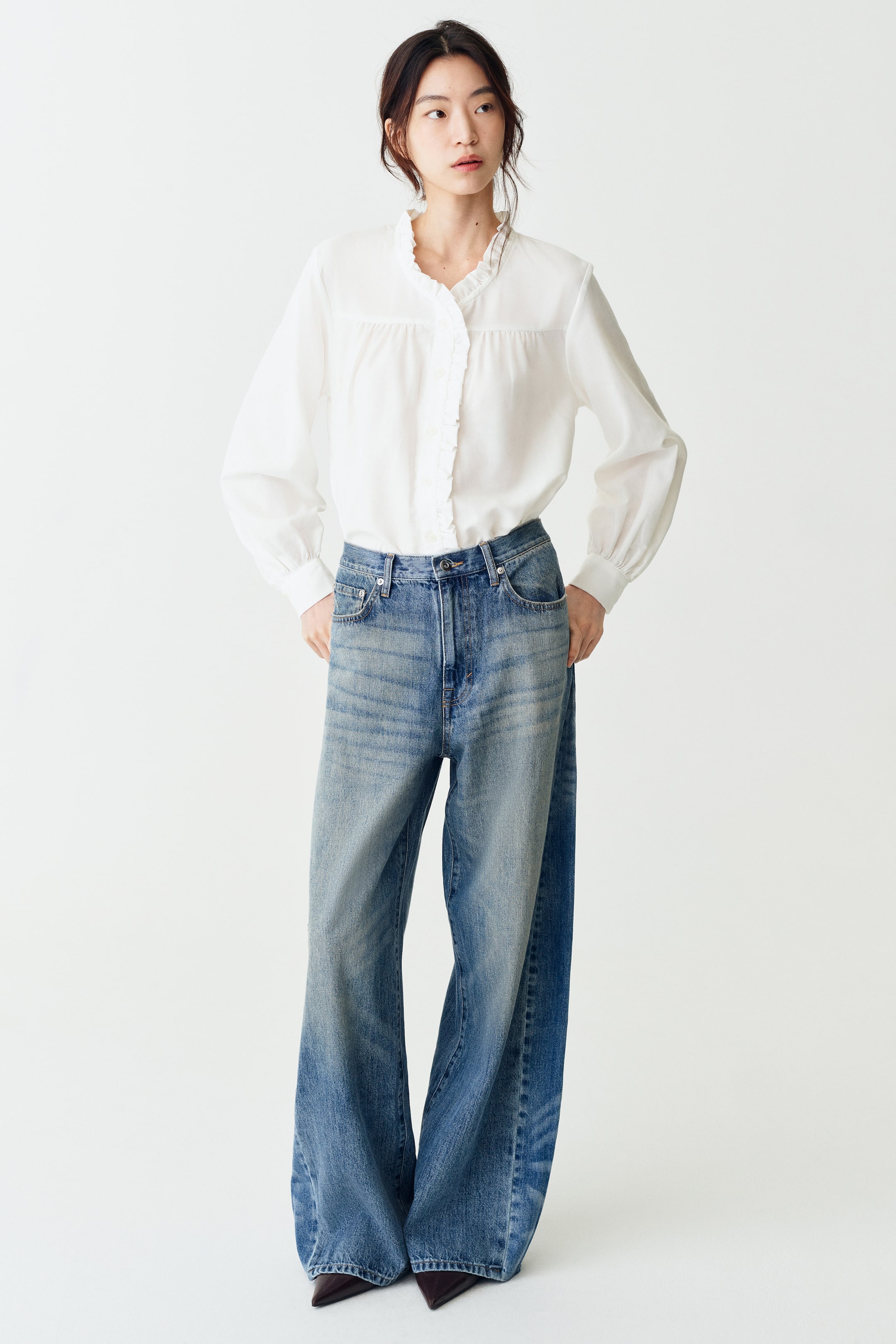 Xem ảnh lớn hơn: Người mẫu đang mặc một chiếc áo blouse twill màu trắng với cổ bèo, đường viền bèo dọc theo nẹp cúc phía trước, phần cầu ngực nhún bèo ở mặt trước và tay áo dài phồng rộng. Áo được sơ vin vào quần jeans xanh ống rộng.