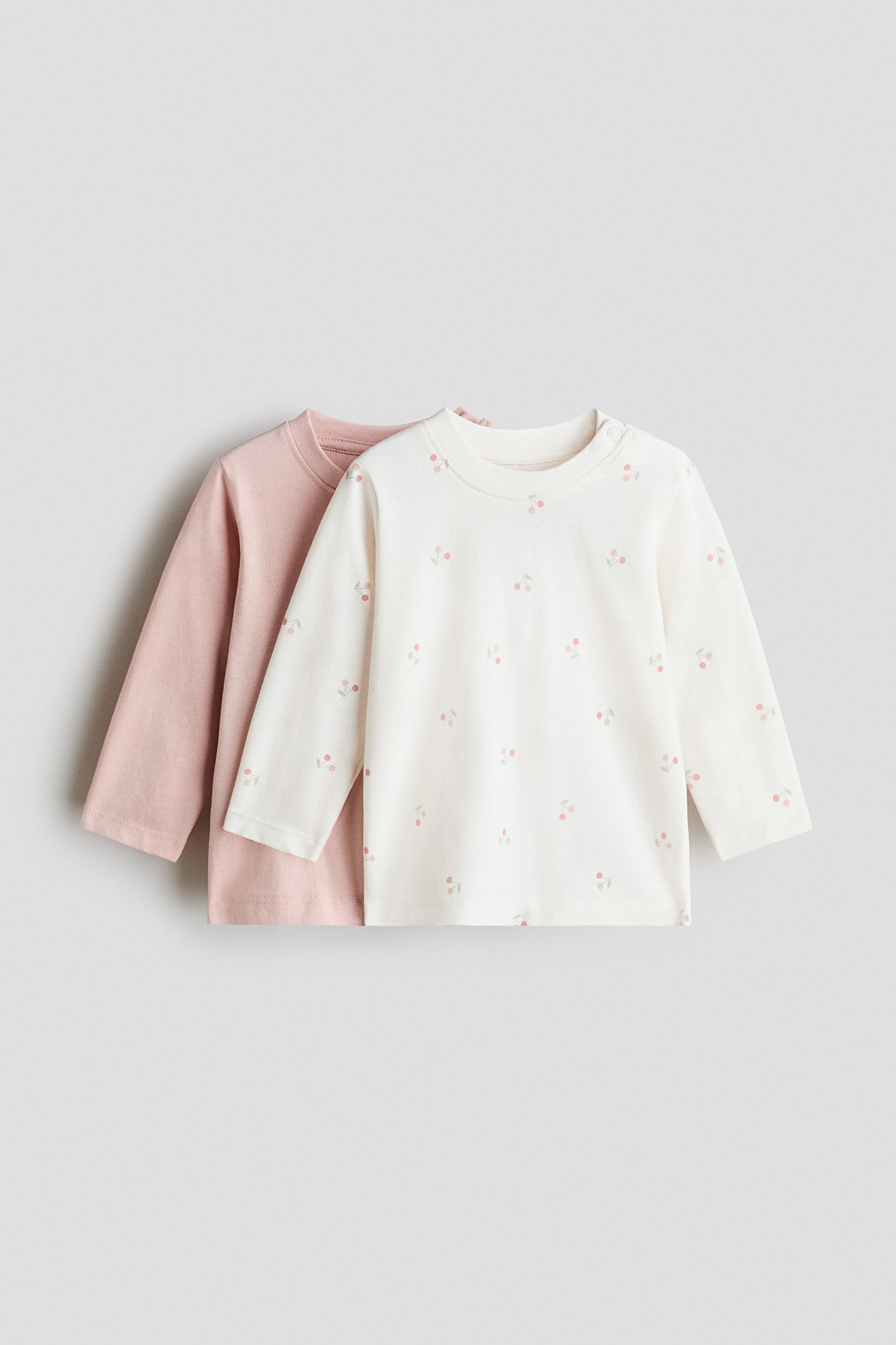 View larger image: This 2-pack of long-sleeved jersey tops features two front-facing tops laid flat and slightly layered. The front top is an off-white jersey with a scattered cherry print and two snap closures on the left shoulder, while the top behind it is a solid light-pink jersey; both feature crew necks and long sleeves.
