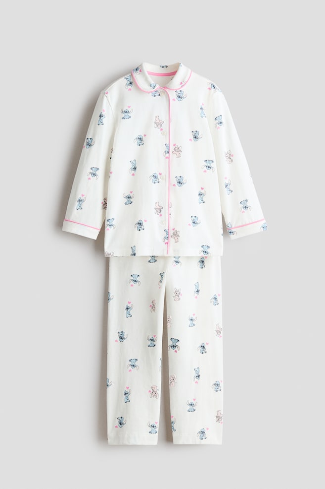 A cream pajama set featuring a repeating pattern of blue Stitch characters, pink Angel characters, and small pink hearts. The long-sleeved top displays a notched collar, a full button placket, and cuffs, all edged with pink piping, while the matching long pants showcase a straight leg design. - 1