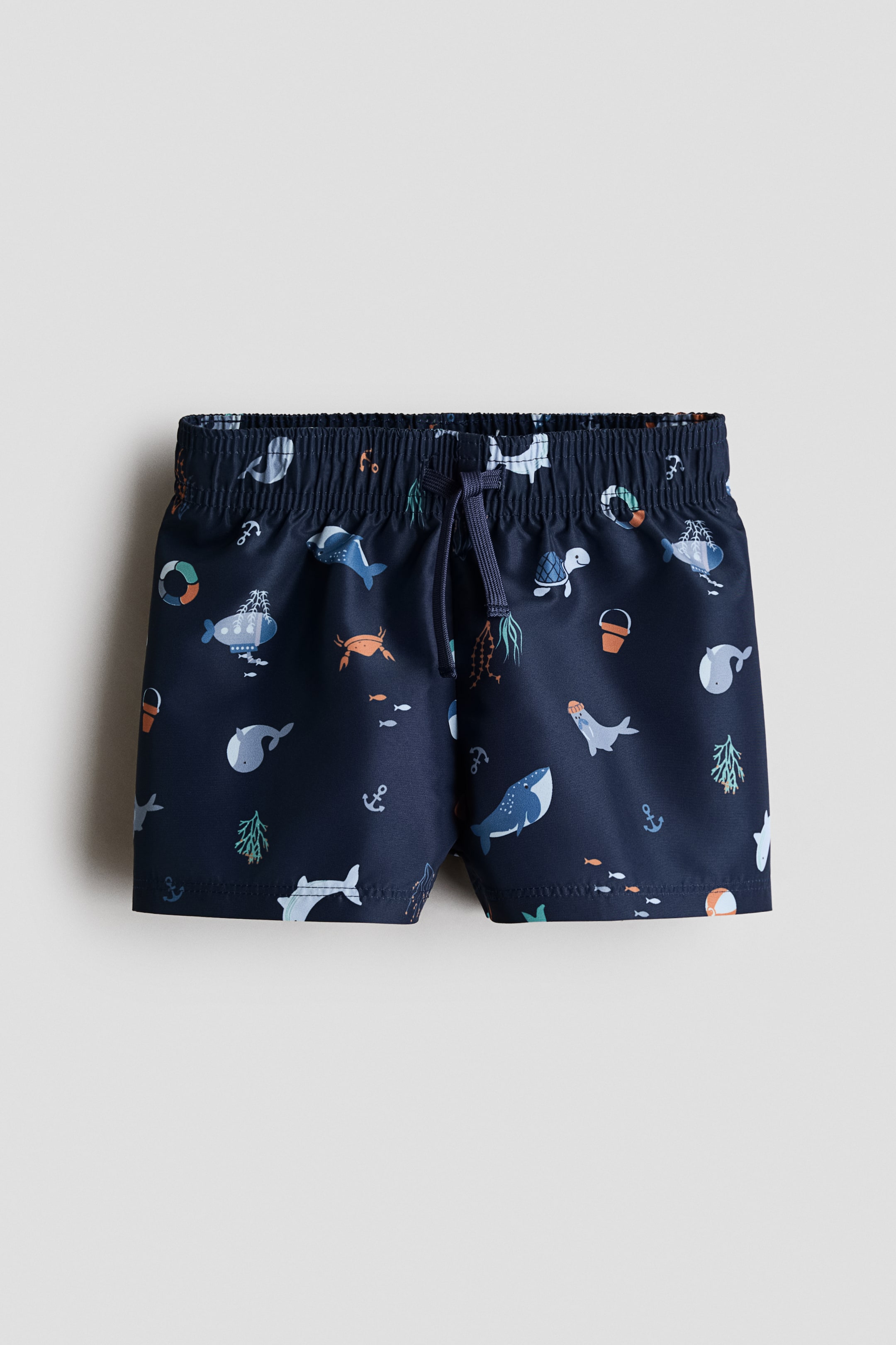 View larger image: Patterned swim shorts are visible from the front, featuring a dark blue base with a multi-coloured marine life design. They are designed with an elasticated drawstring waist, with the drawstring tied at the centre.
