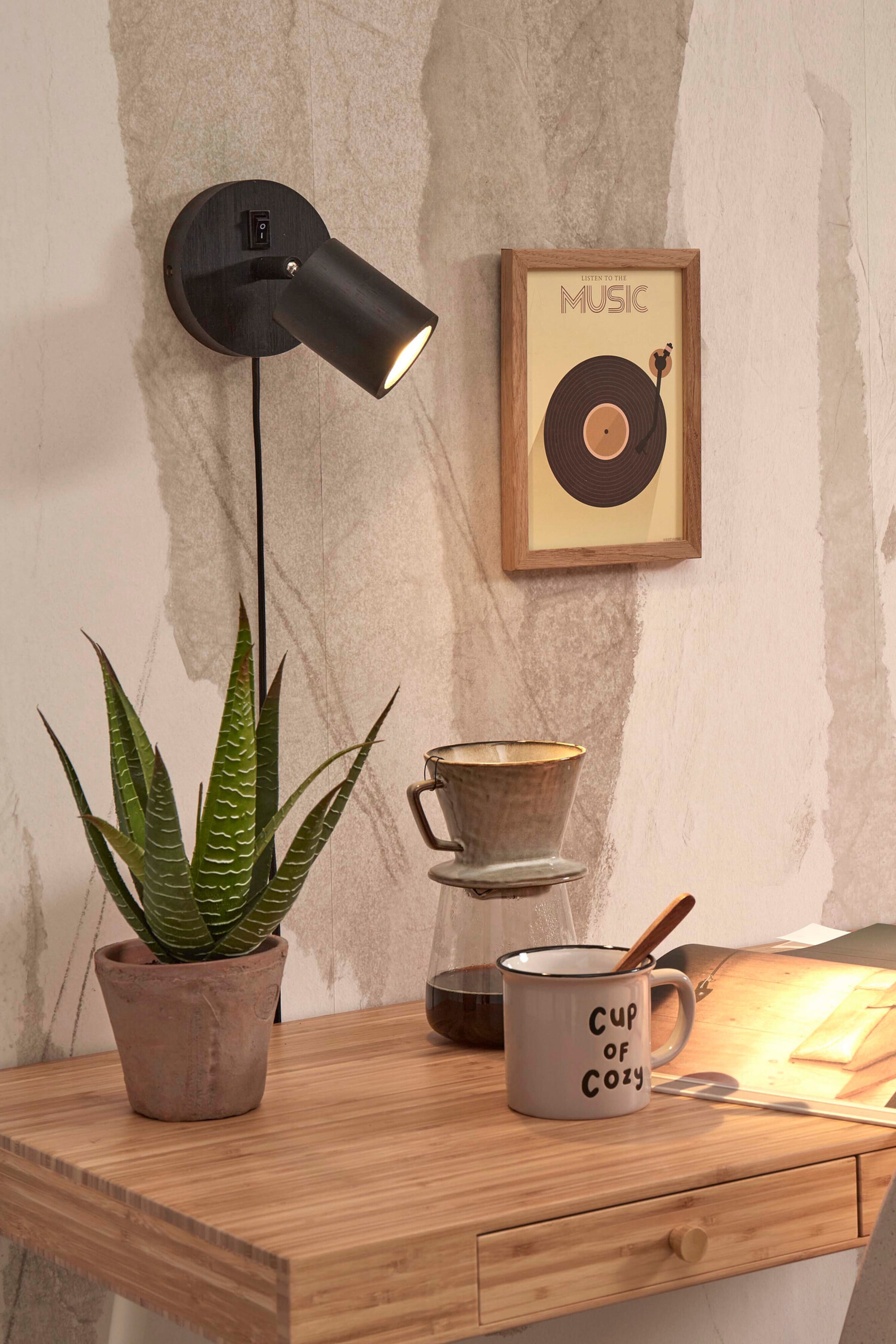 Agrandir l'image: Un spot noir mural est présenté, doté d’une base circulaire en bois avec un interrupteur à bascule et une tête de lampe cylindrique orientable. La tête de lampe est inclinée vers le bas et diffuse une lumière chaude, tandis qu’un fin cordon noir pend verticalement sous la base.