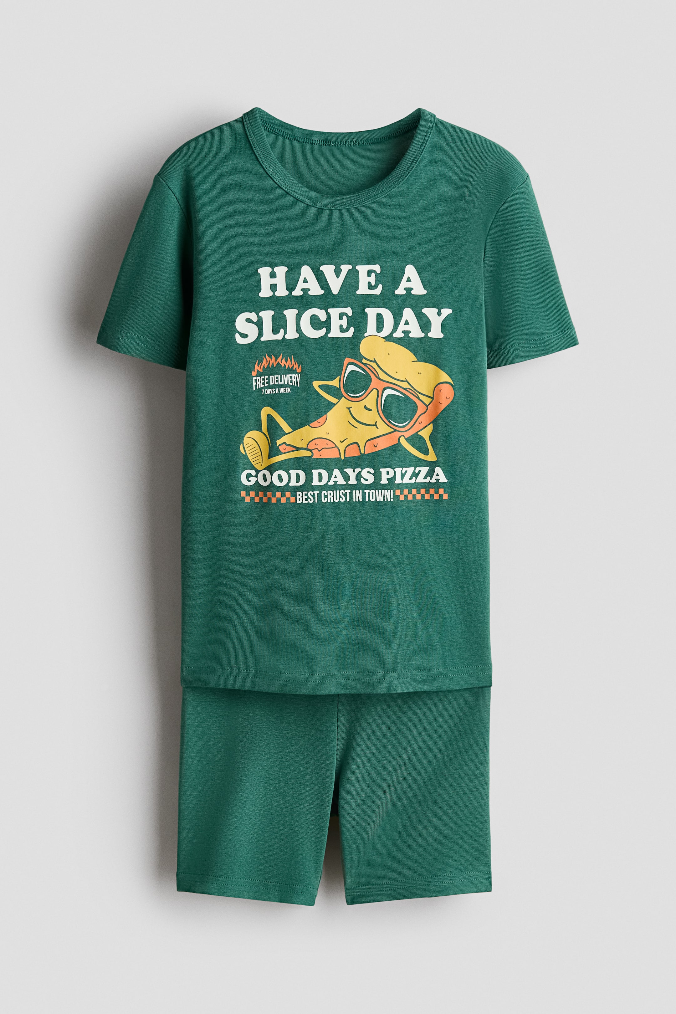 View larger image: A pajama set includes a T-shirt and a pair of shorts. The T-shirt, shown from the front, is green and features a print design on the front with the text "HAVE A SLICE DAY" and an illustration of a smiling pizza slice wearing sunglasses. The matching green shorts have an elastic waistband.
